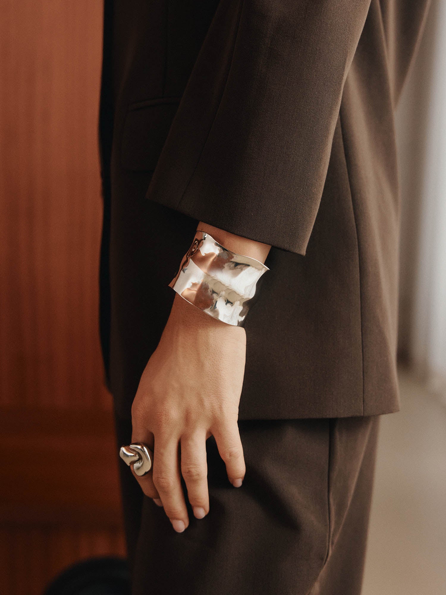 Eine Person in einem braunen Anzug steht mit dem Arm an der Seite und hebt die LeGer x Purelei - Modern Muse Armfessel von LeGer by Lena Gercke und einen großen silbernen Ring hervor, wobei der Fokus auf dem Edelstahlschmuck und der Hand vor einem sanft verschwommenen Hintergrund liegt.