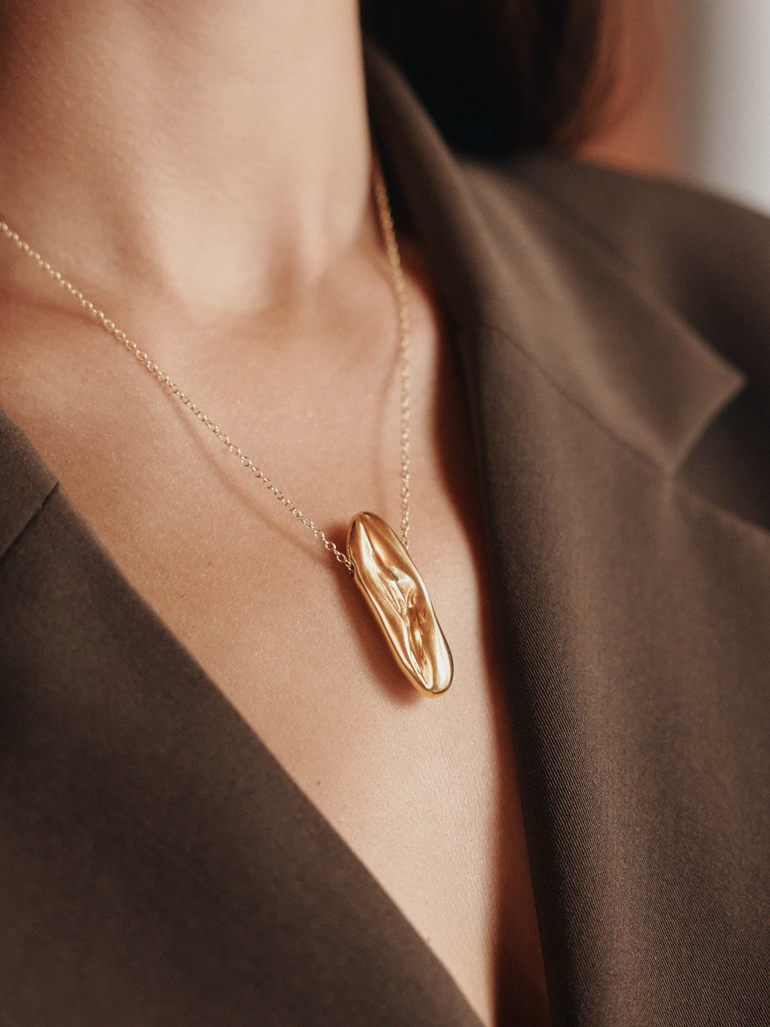 Eine Person in einem braunen Blazer präsentiert die LeGer x Purelei - Modern Muse Kette von LeGer by Lena Gercke, die einen abstrakten Goldanhänger aufweist. Der Blick fällt auf den eleganten Ausschnitt und die stilvolle Edelstahlkette.