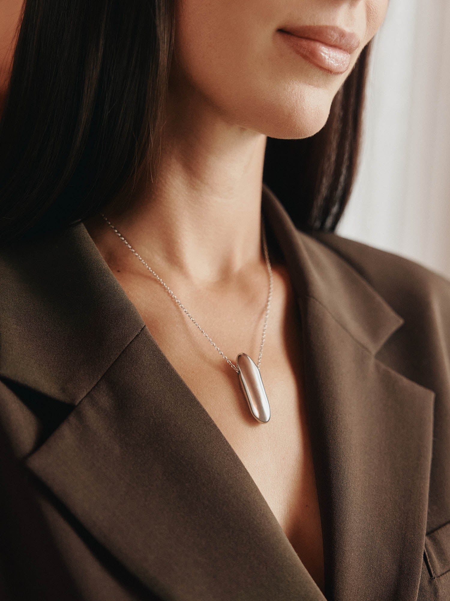 Eine Frau trägt einen dunkelbraunen Blazer und die LeGer x Purelei - Modern Muse Kette von LeGer by Lena Gercke, eine auffällige ovale Silberkette mit Anhänger. Das Bild hebt ihr unteres Gesicht, den Hals und die obere Brust hervor.