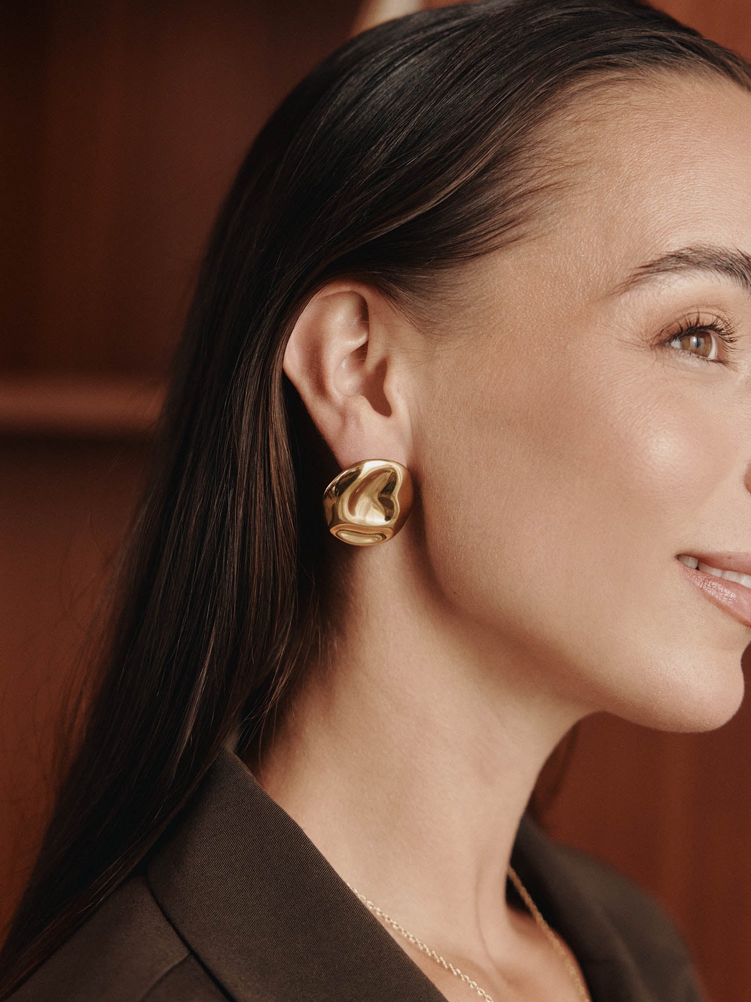 Eine Frau mit glattem dunklem Haar trägt LeGer x Purelei - Modern Muse Ohrringe von LeGer by Lena Gercke, gestylt mit einem braunen Blazer. Sie lächelt sanft in einer Nahaufnahme im Seitenprofil vor einem verschwommenen braunen Hintergrund - ein echtes Statement.