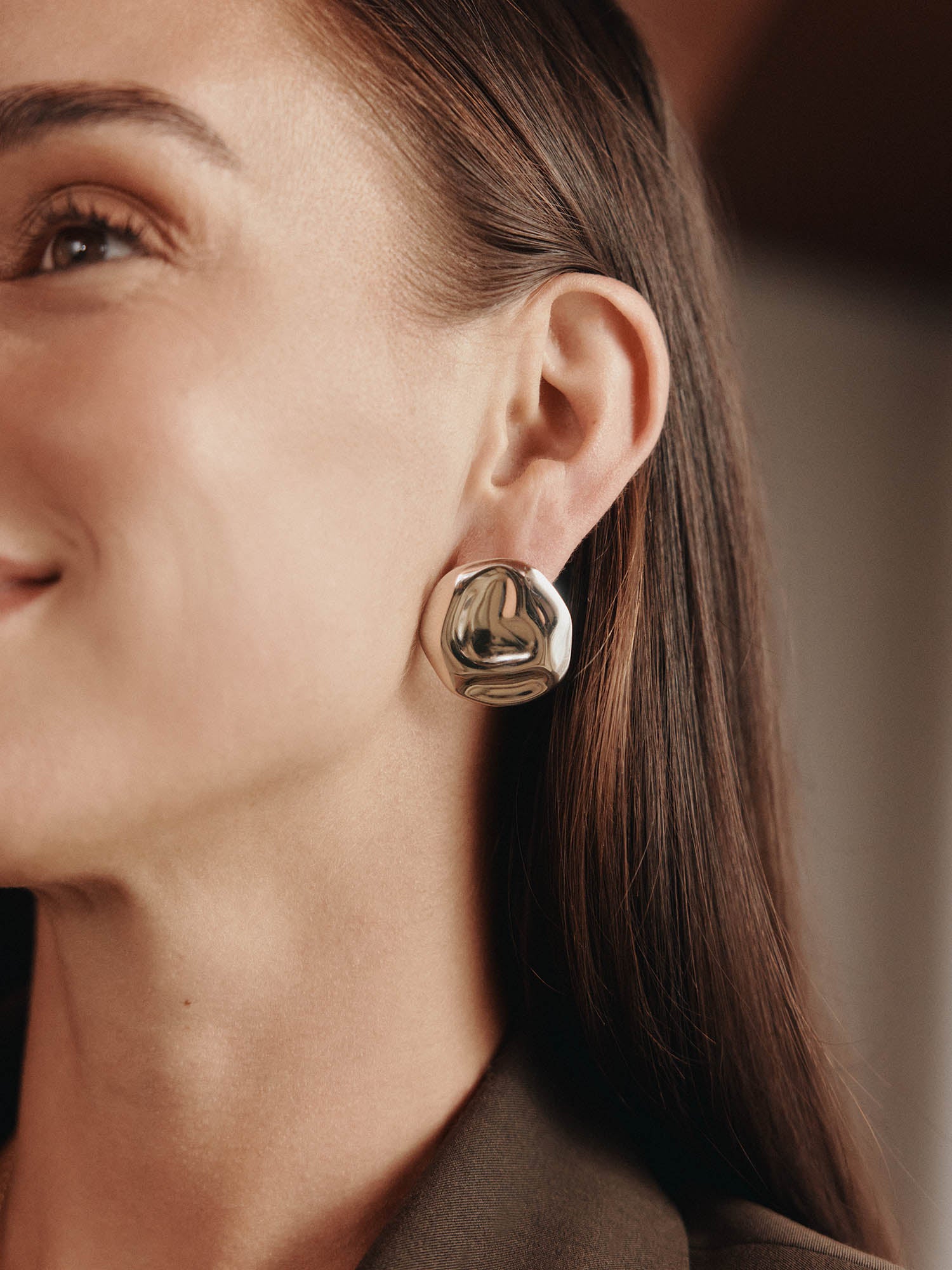 Eine Nahaufnahme einer Frau mit glattem braunem Haar, die glänzende, abstrakt geformte LeGer x Purelei - Modern Muse Ohrringe von LeGer by Lena Gercke trägt. Sie lächelt leicht vor einem sanft verschwommenen Hintergrund.