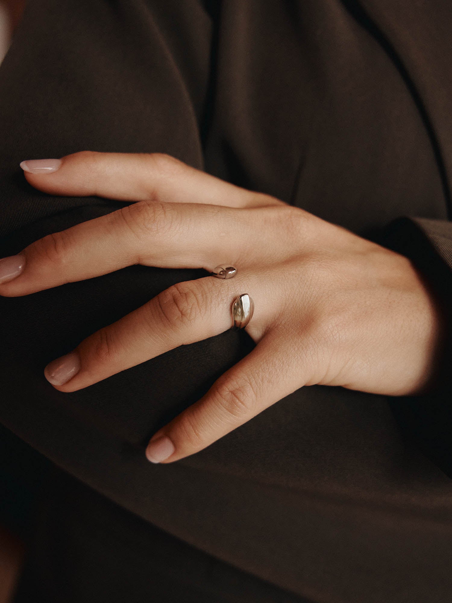 Die Nahaufnahme einer ordentlich manikürten Hand zeigt den LeGer x Purelei - Open Mind Ring von LeGer by Lena Gercke am Mittelfinger, der sanft auf einem dunkelbraunen Stoffhintergrund ruht.