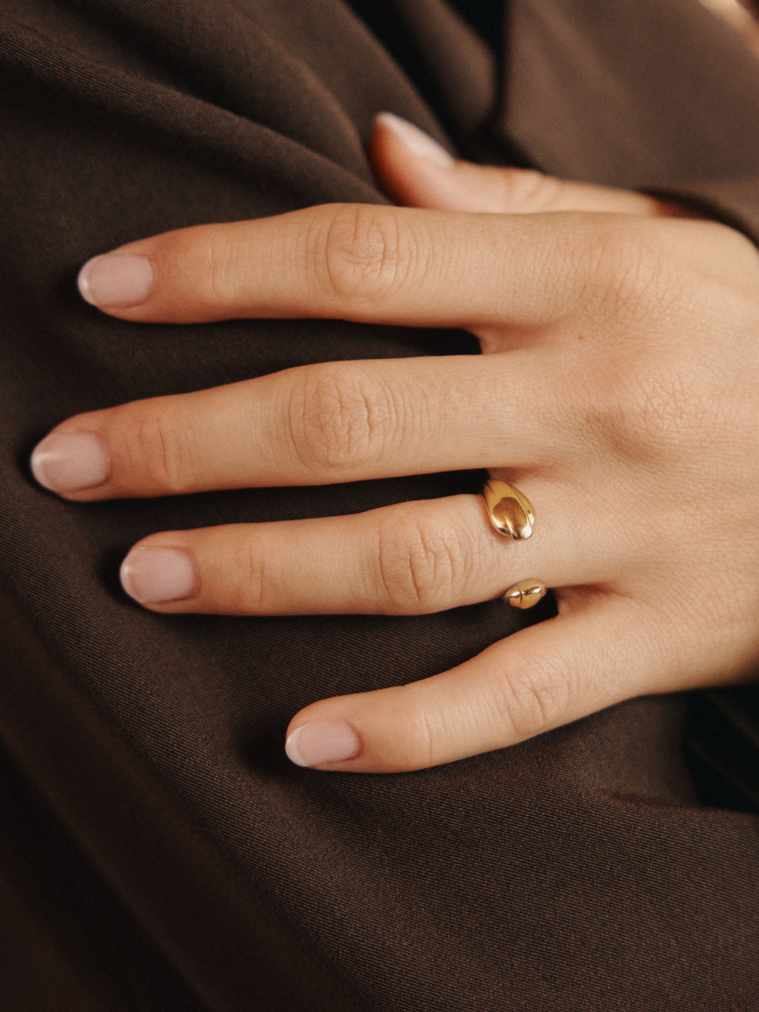 Nahaufnahme einer Hand mit hellrosa manikürten Nägeln, die den LeGer x Purelei - Open Mind Ring von LeGer by Lena Gercke in Gold am Ringfinger trägt, der auf braunem Stoff ruht.