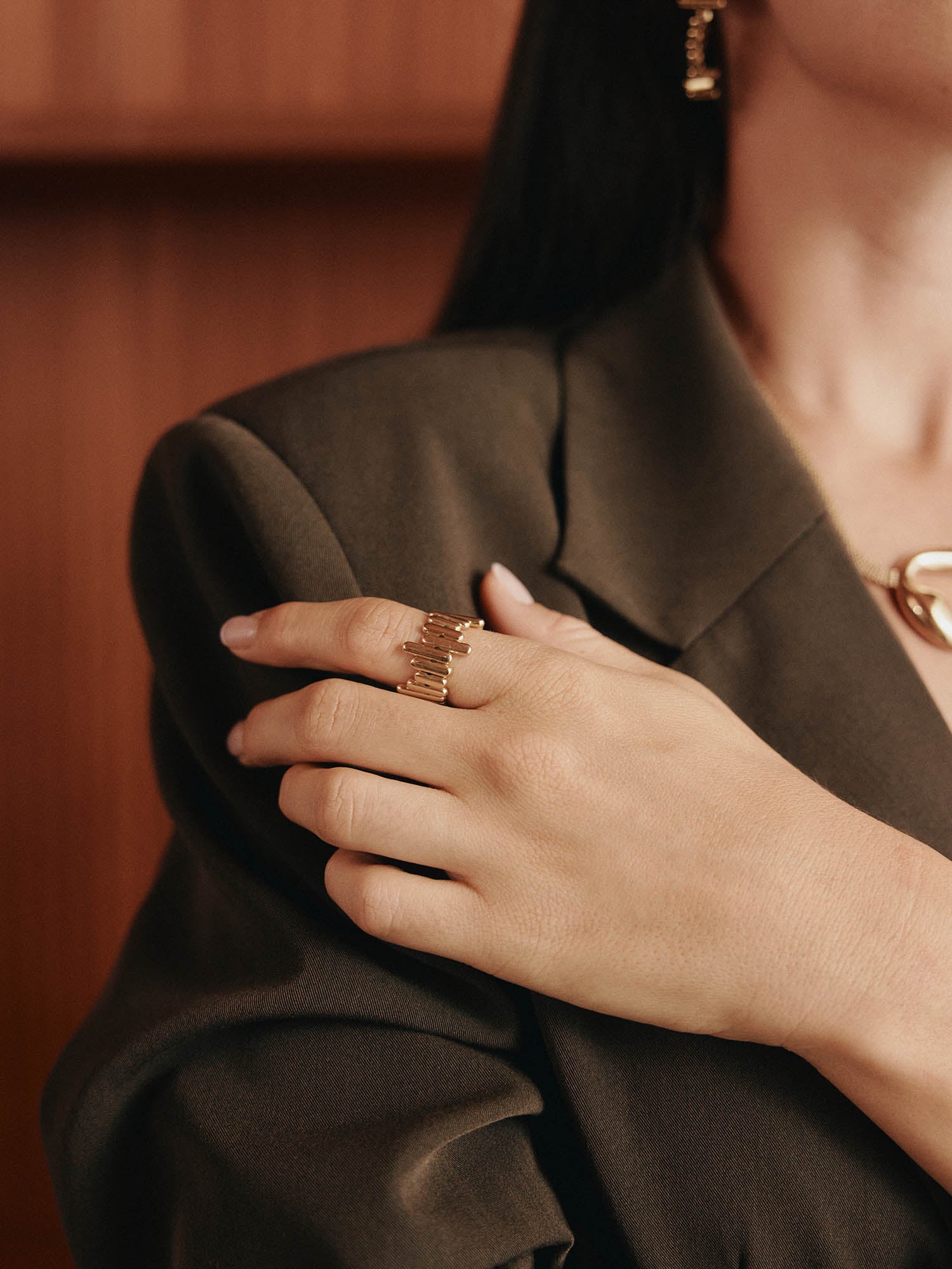 Eine Person in einem braunen Blazer berührt ihre Brust und hebt den LeGer x Purelei - Rise Ring von LeGer by Lena Gercke und einen passenden goldenen Ohrring hervor. Der Schmuck und der strukturierte Blazer sind der Hauptfokus.