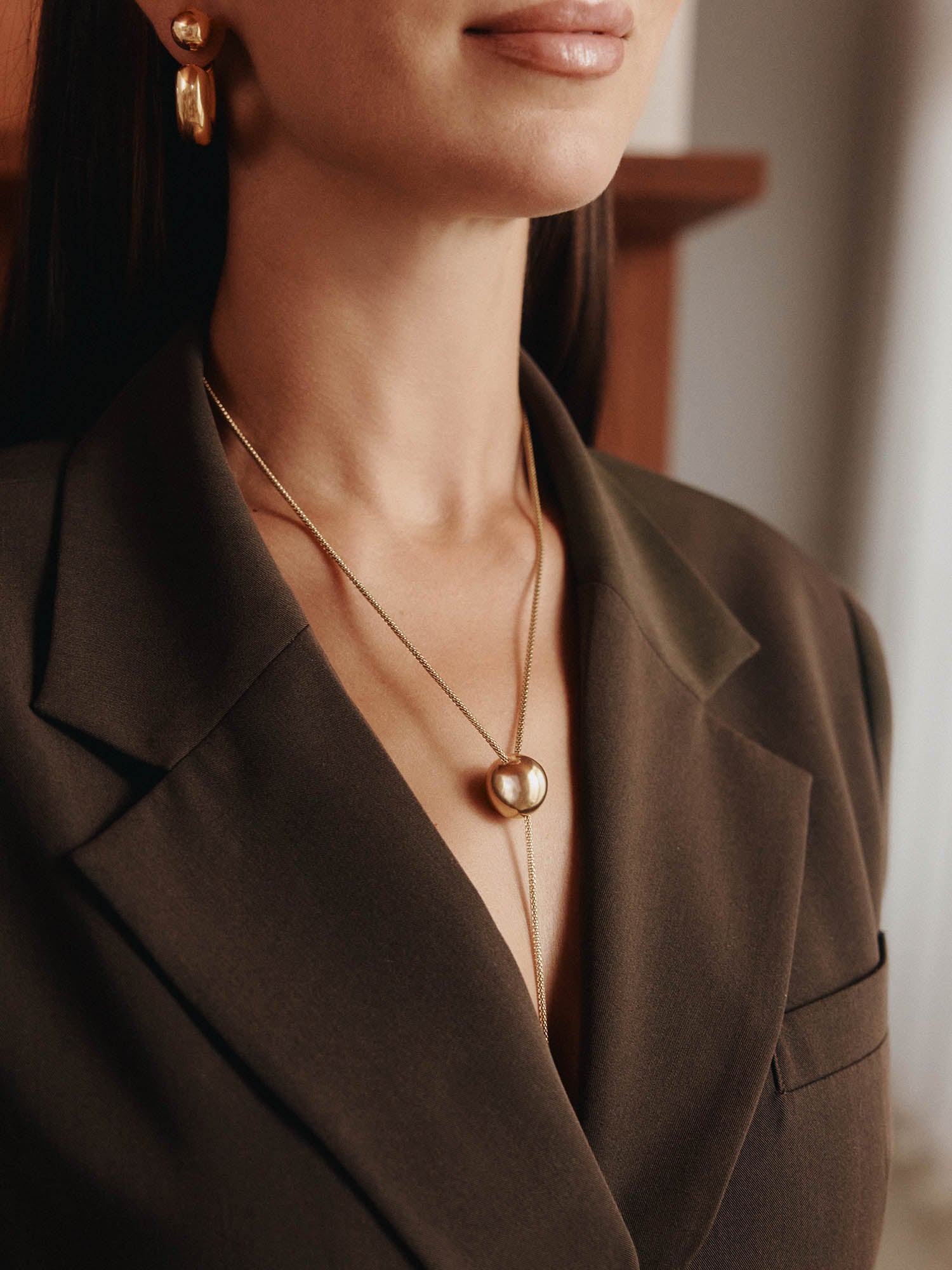 Eine Frau in einem dunklen Blazer, goldenen Ohrringen und zwei goldenen Lagenketten - darunter die LeGer x Purelei - Unstoppable Kette von LeGer by Lena Gercke - wird von den Schultern bis knapp über den Mund gezeigt.