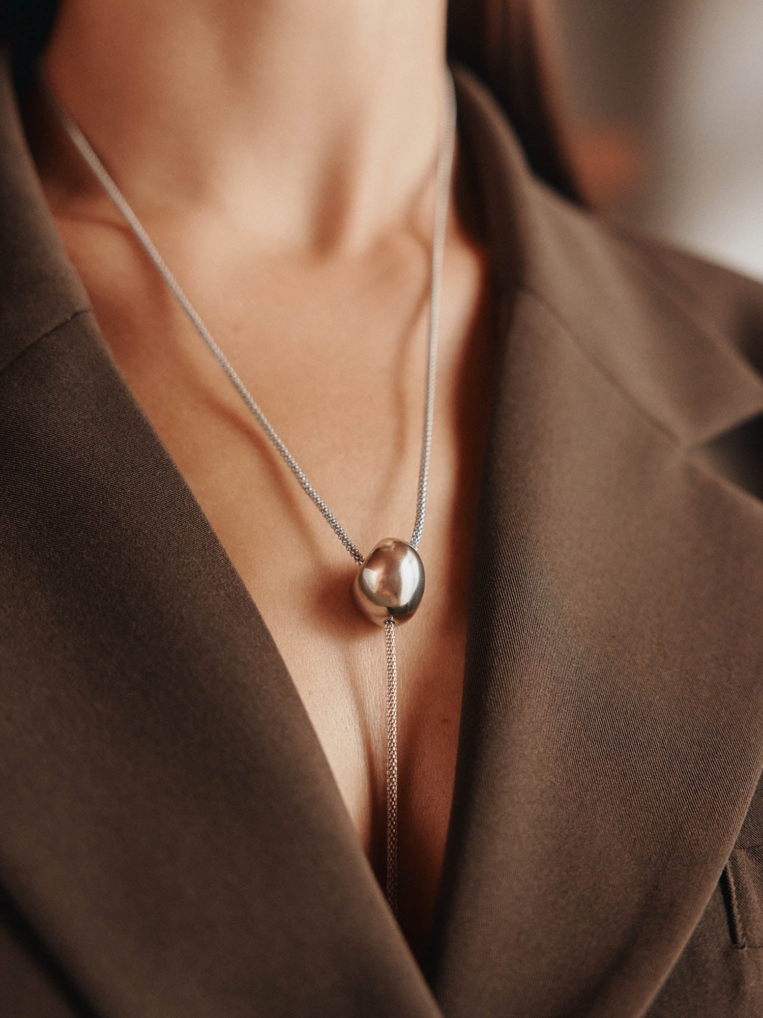 Eine Nahaufnahme zeigt eine Person in einem braunen Blazer mit tiefem Ausschnitt, die die LeGer x Purelei - Unstoppable Kette von LeGer by Lena Gercke trägt - eine silberne Halskette mit einem glatten, runden Anhänger, der auf ihrer Brust ruht.