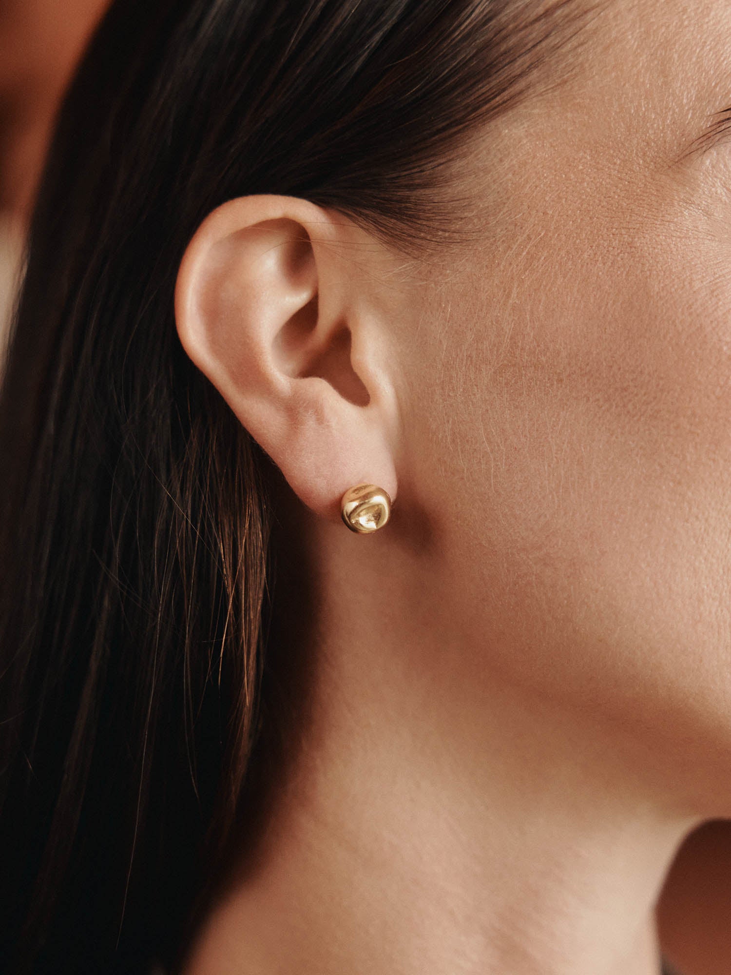 Nahaufnahme des Ohrs einer Frau, die LeGer x Purelei - Unstoppable Ohrringe von LeGer by Lena Gercke, einen kleinen runden goldenen Ohrstecker, trägt. Ihr glattes braunes Haar ist hinter ihr Ohr gesteckt, und ihre glatte Haut ist sanft beleuchtet.
