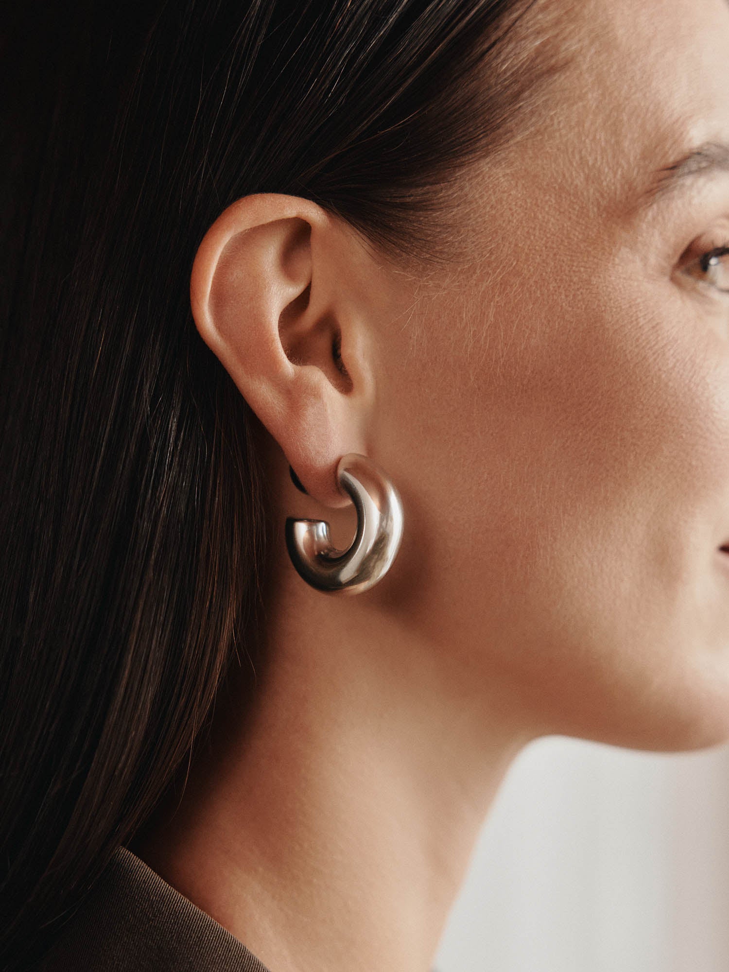 Seitenansicht einer Frau mit glattem braunem Haar in einem braunen Outfit, die LeGer x Purelei - Unstoppable Ohrringe von LeGer by Lena Gercke trägt und deren schickes 4-in-1-Design präsentiert. Ihre glatte Haut und der sanft verschwommene Hintergrund verleihen ihr einen eleganten Touch.