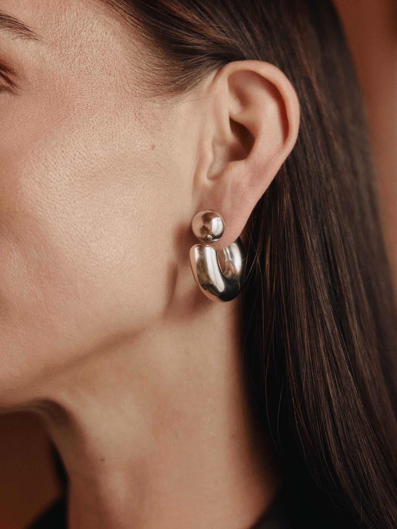 Nahaufnahme des Ohrs einer Frau, die LeGer x Purelei - Unstoppable Ohrringe by LeGer by Lena Gercke - glänzende Silberohrringe mit einem runden Ohrstecker und einem dicken, geschwungenen Reif trägt. Ihr glattes braunes Haar hat sie ordentlich hinter dem Ohr versteckt.