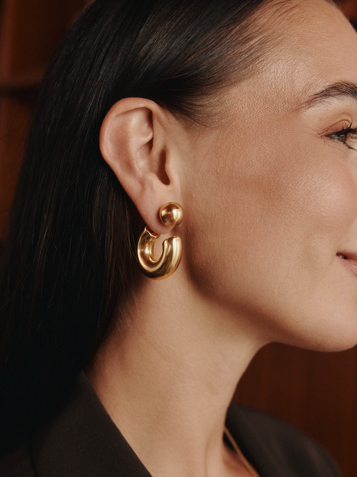Nahaufnahme einer Frau im Profil mit langen dunklen Haaren, die die LeGer x Purelei - Unstoppable Ohrringe von LeGer by Lena Gercke - glänzende, klobige Goldreifen trägt. Der sanft verschwommene Hintergrund leuchtet in warmen Brauntönen.