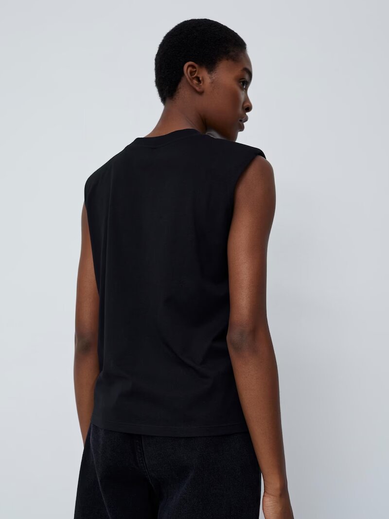 Eine Person, die von der Kamera abgewandt ist, trägt ein Top „Kim“ von LeGer by Lena Gercke, ein schwarzes ärmelloses Design mit normaler Länge, kombiniert mit einer dunklen Hose vor einem schlichten hellen Hintergrund.