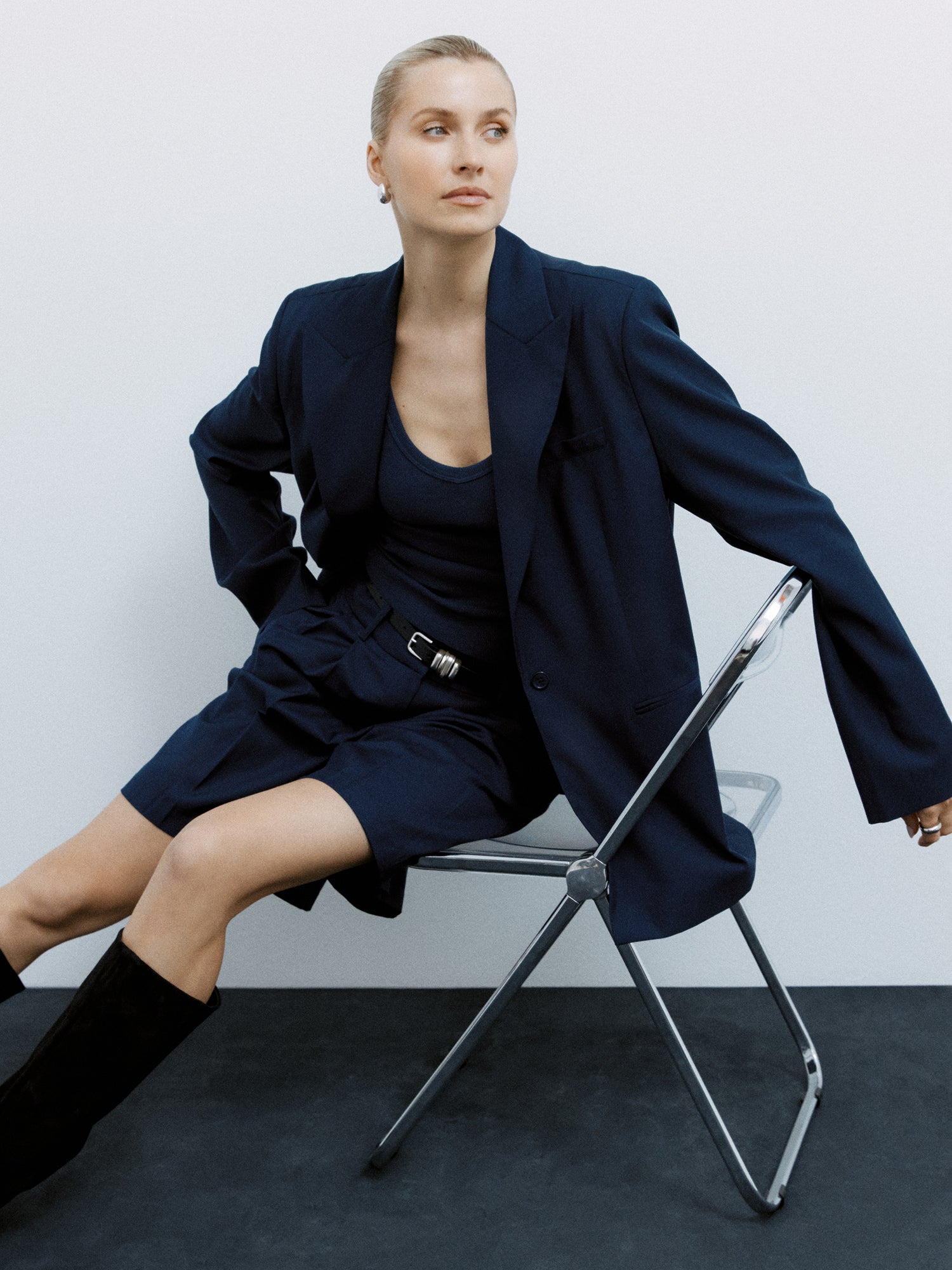 Eine Person mit zurückgebundenen Haaren sitzt stylisch auf einem Metallstuhl. Sie trägt einen dunklen Blazer über einem passenden Oberteil und Dita Shorts von LeGer by Lena Gercke, die mit einem Gürtel zusammengehalten werden. Kniehohe schwarze Stiefel runden das Ensemble ab. Ein Arm ruht auf der Stuhllehne und strahlt vor dem schlichten Hintergrund mühelosen Stil aus.