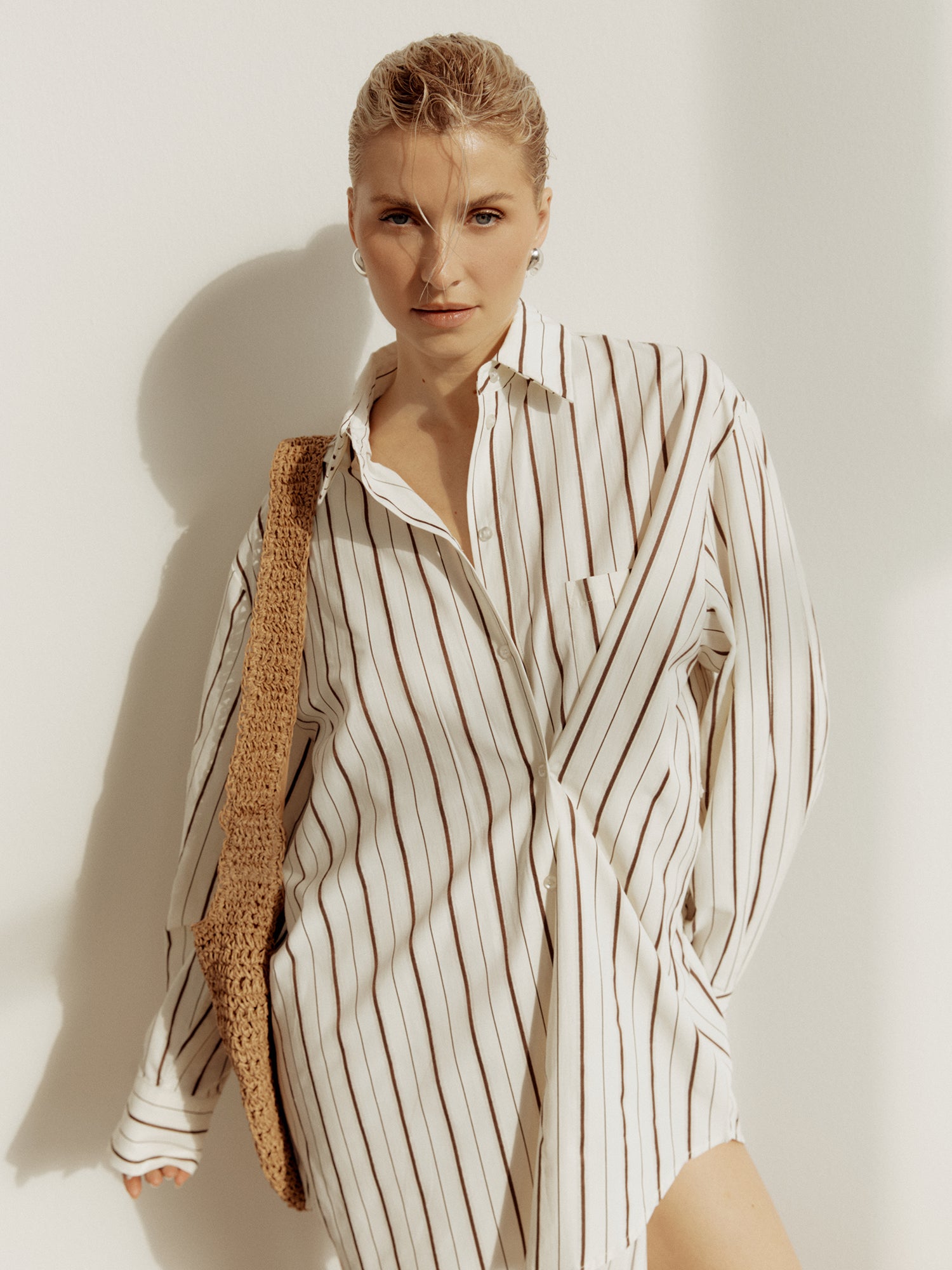 Eine Person mit kurzen blonden Haaren trägt das Ela Dress von LeGer by Lena Gercke und eine gewebte Umhängetasche und steht vor einer sonnenbeschienenen weißen Wand.