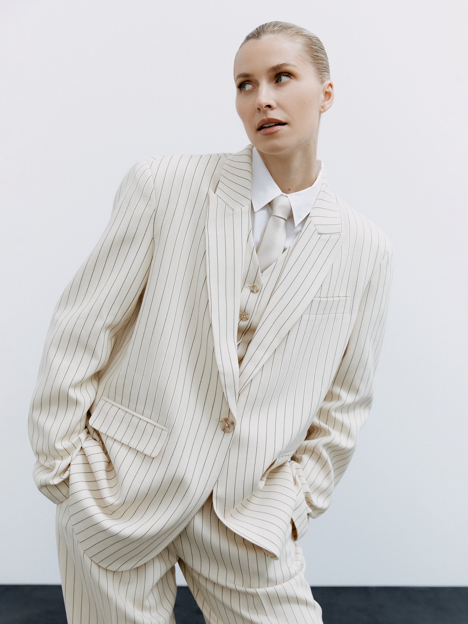 Eine Person mit blondem, nach hinten gebundenem Haar präsentiert den übergroßen, cremefarbenen Nadelstreifen-Blazer „Elaine“ von LeGer by Lena Gercke, kombiniert mit einer passenden Weste und einem weißen Hemd. Die Hände stecken in den Taschen, während sie vor einem schlichten weißen Hintergrund steht.