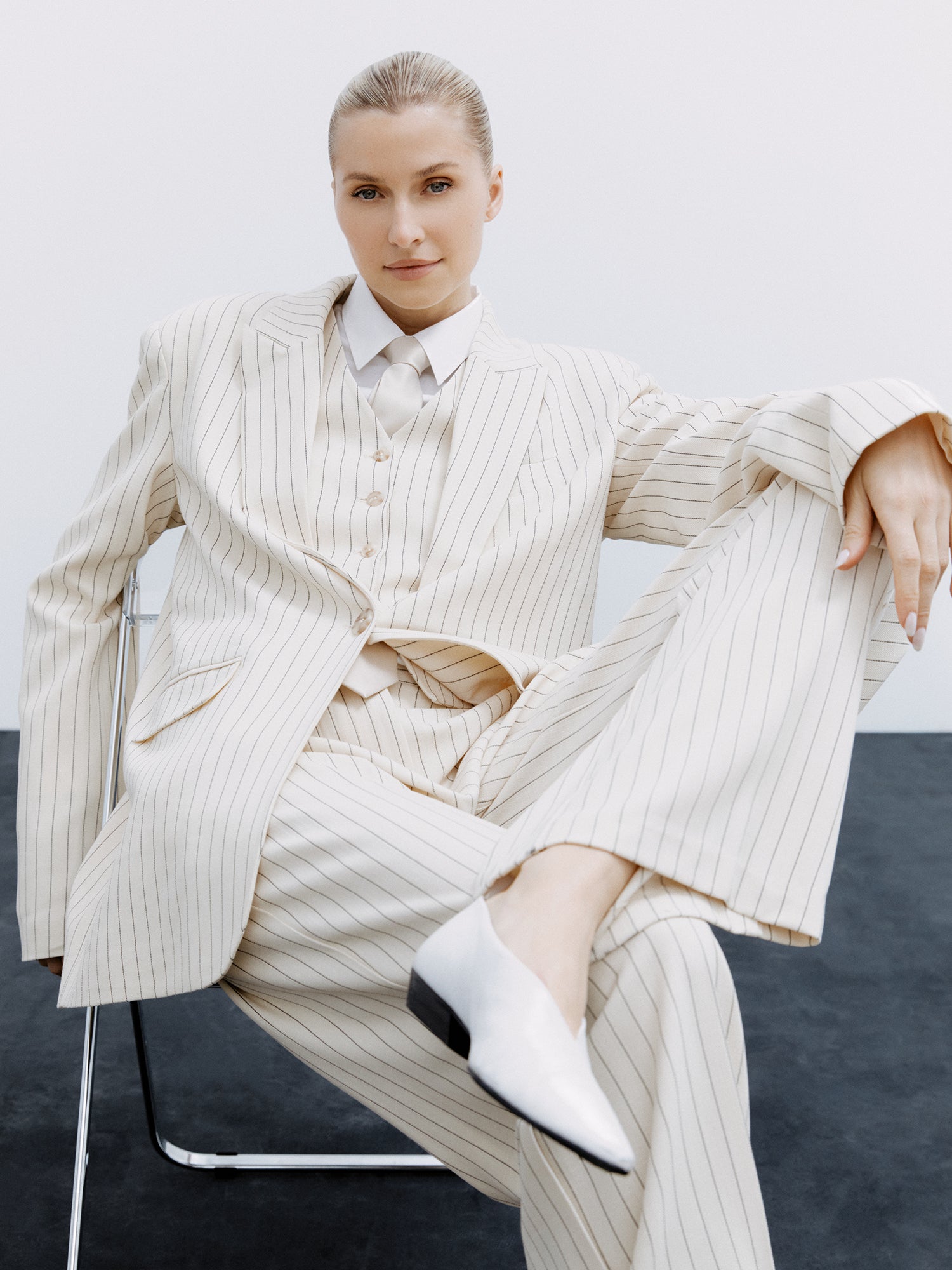 Eine Person in cremefarbenem Nadelstreifenanzug und weißem Hemd sitzt lässig mit übereinandergeschlagenen Beinen auf einem Stuhl und strahlt lässigen Stil aus. Ihr zurückgekämmtes Haar harmoniert perfekt mit der Elaine-Weste von LeGer by Lena Gercke und den weißen Schuhen vor dem minimalistischen Kontrast einer schlichten weißen Wand und eines dunklen Bodens.