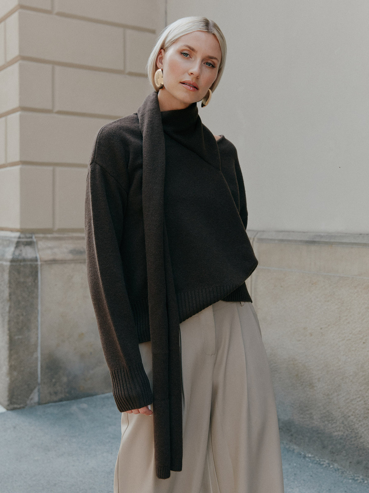Eine blonde Frau trägt den Pullover „Helga“ von LeGer by Lena Gercke, einen locker sitzenden, dunkelbraunen Rollkragenpullover, dazu einen langen Schal, eine hellbeige Hose mit weitem Bein und große goldene Ohrringe.