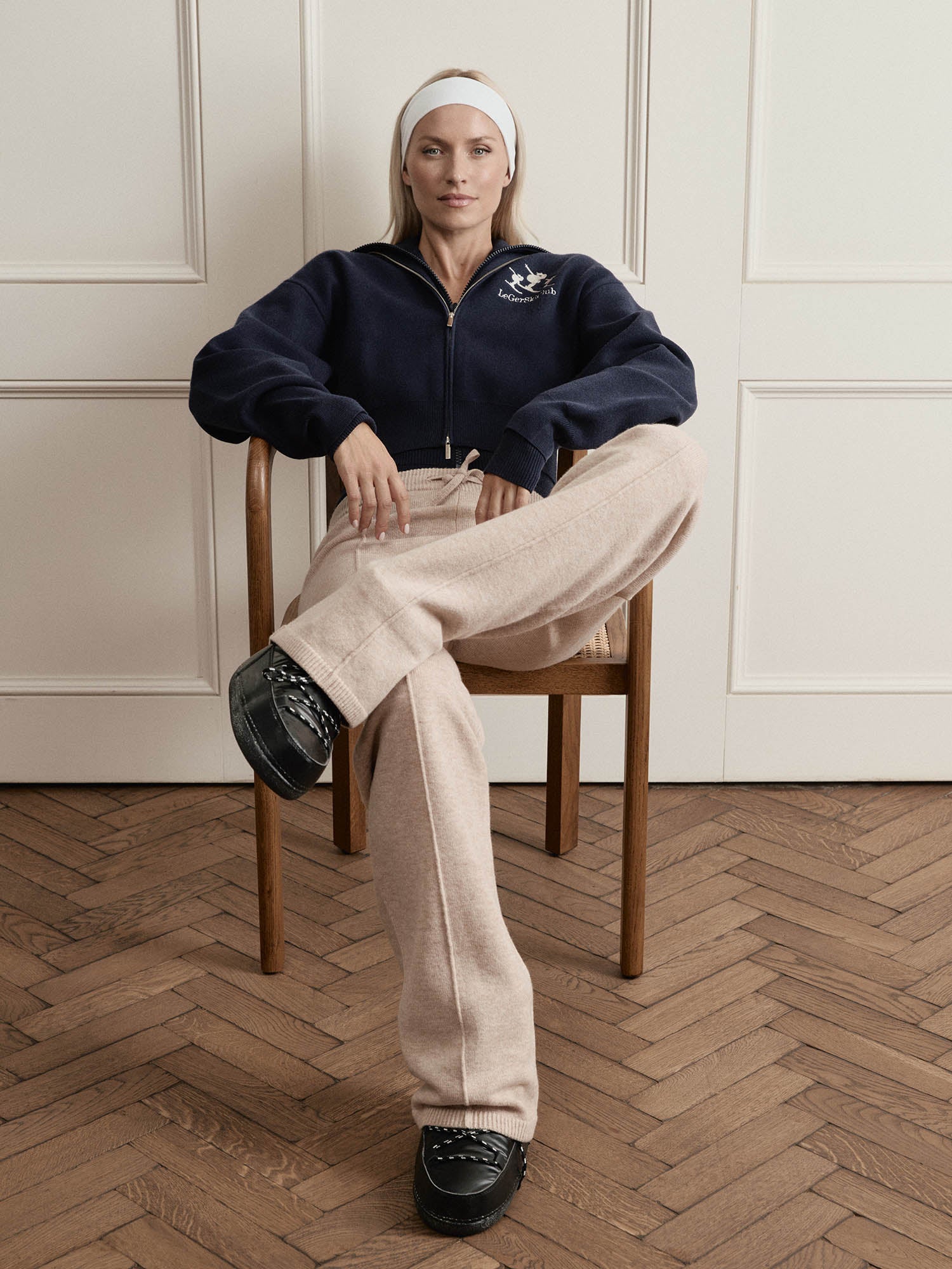 Eine Frau, die den navyfarbenen Jaden-Pullover von LeGer by Lena Gercke mit extra langen Ärmeln in Größe 36 trägt, sitzt selbstbewusst auf einem Holzstuhl mit beiger Hose, schwarzen Schuhen und weißem Stirnband, die Arme auf die Stuhllehne gestützt.