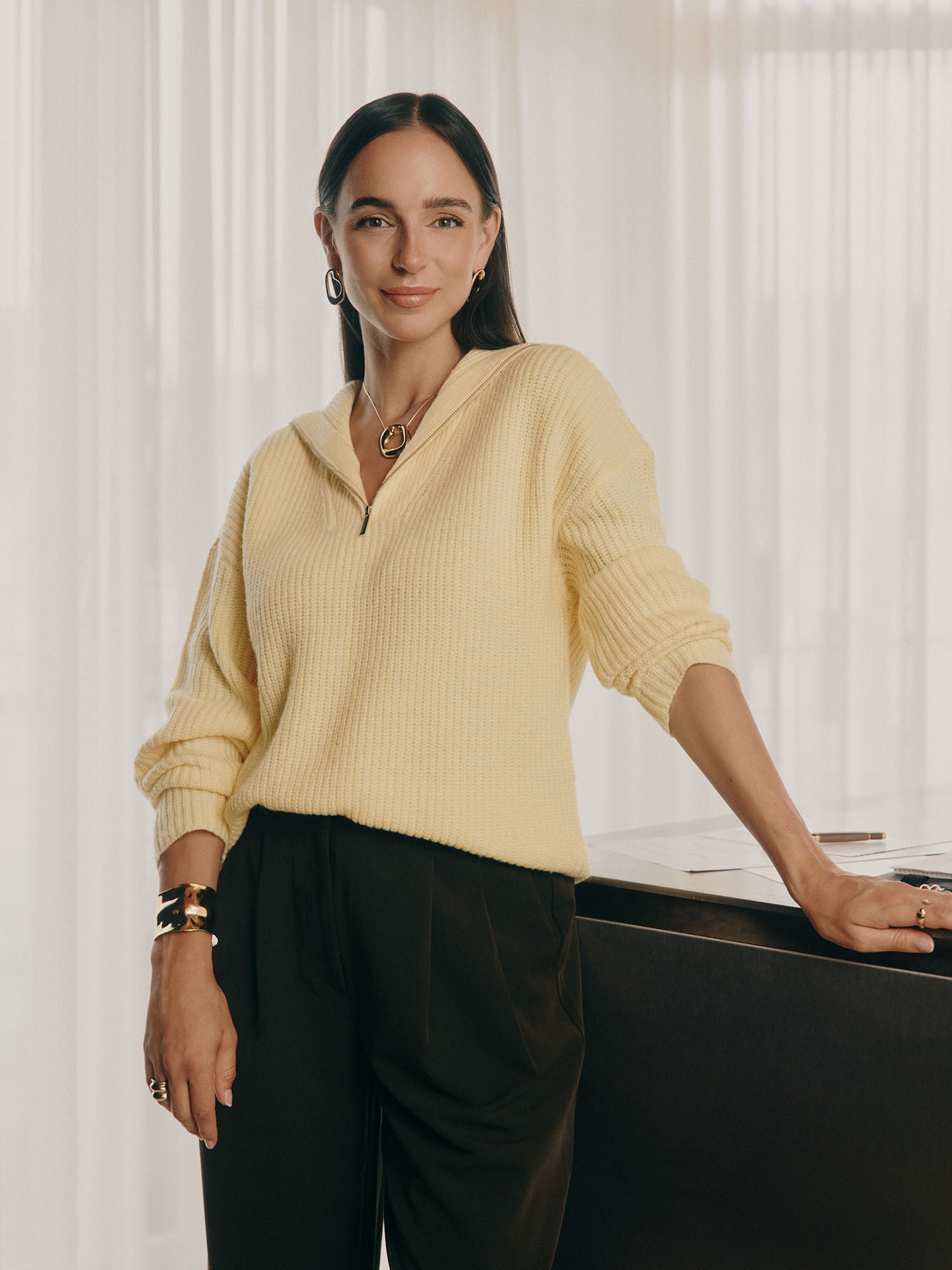 Eine Frau mit langen dunklen Haaren lächelt in einem Innenraum. Sie trägt den LeGer x Purelei - Lea Jumper by LeGer by Lena Gercke - einen hellgelben Strickpullover mit extralangen Ärmeln - und eine schwarze Hose, die mit Goldschmuck verziert ist. Im Hintergrund sind helle Vorhänge und ein dunkler Tisch zu sehen.