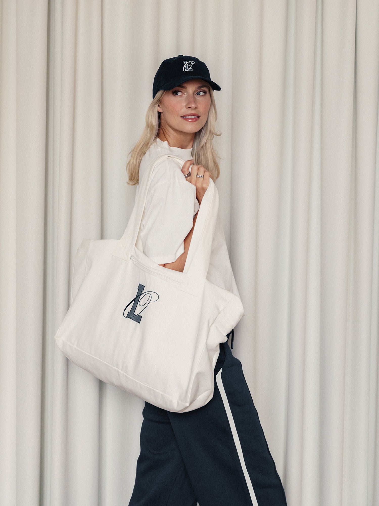 Eine blonde Frau (1,73 m groß, Größe 36) lächelt vor beigen Vorhängen, trägt eine schwarze Mütze, ein weißes T-Shirt, eine dunkle Hose mit weißem Streifen und hat den LeGer x Oh April - Anni Shopper von LeGer by Lena Gercke dabei - eine große weiße Tasche mit Line-Art-Design.