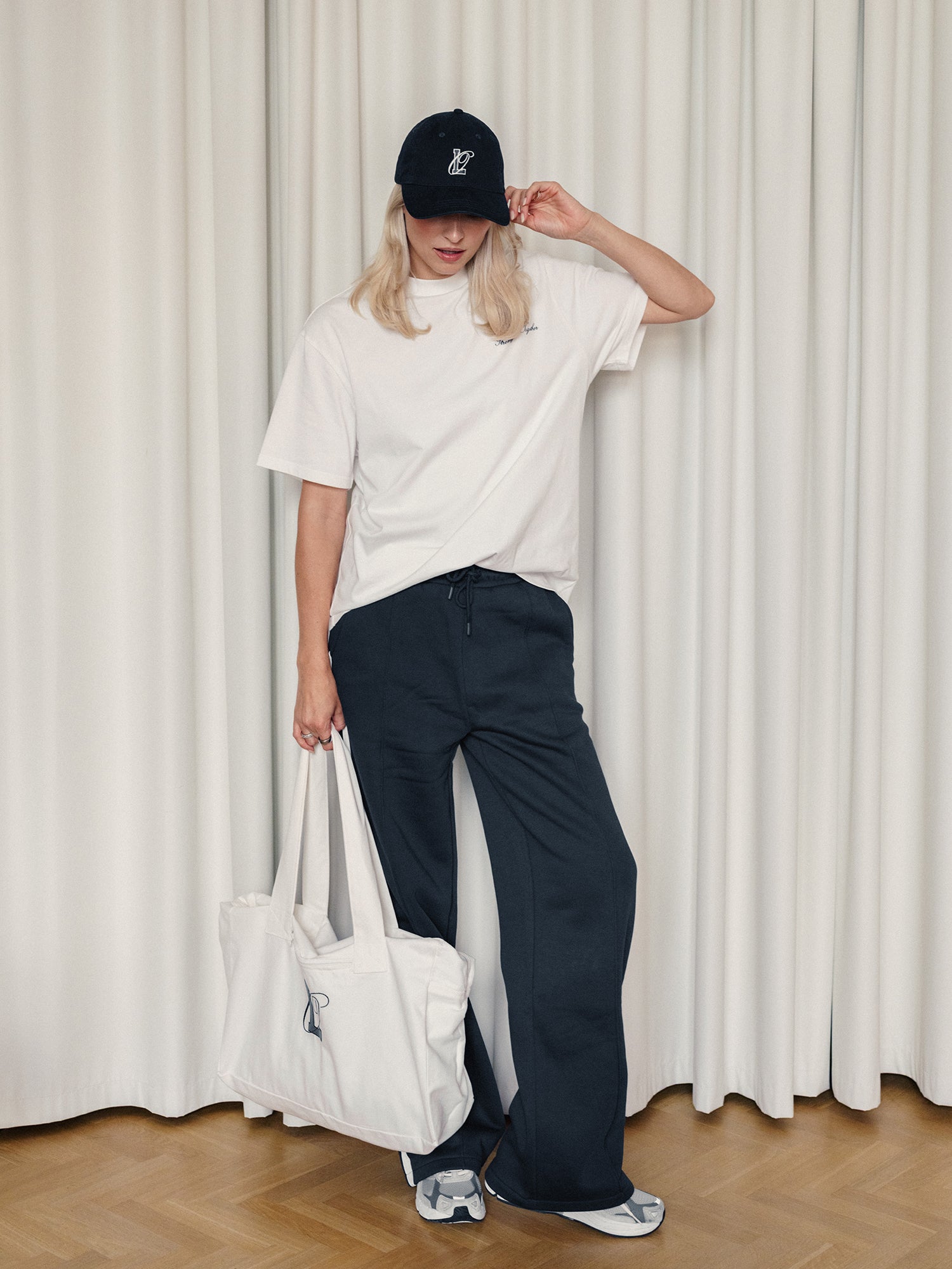 Eine Person trägt die LeGer x Oh April - Arina Cap Stronger von LeGer by Lena Gercke mit einem weißen T-Shirt, einer marineblauen Hose mit weitem Bein, Turnschuhen und hat eine große weiße Tasche dabei. Das Model ist 1,73 m groß, trägt Größe 36 und posiert in einem Raum vor weißen Vorhängen auf einem Holzfußboden.