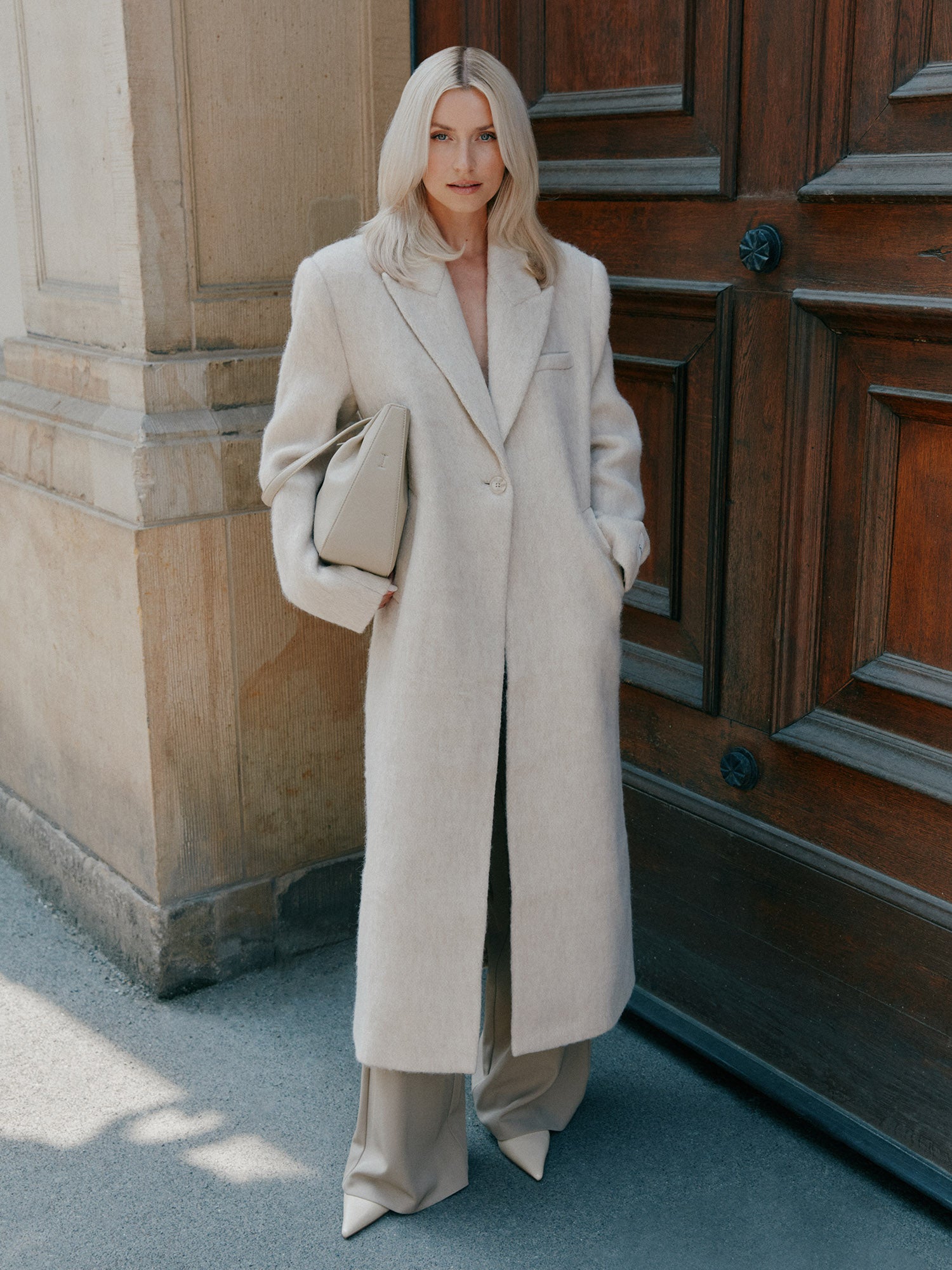 Eine Person mit langen blonden Haaren steht an einer Holztür. Sie trägt den Linda Mantel von LeGer by Lena Gercke in eleganter Regular Fit Passform über einer weiten Hose und hält eine passende beigefarbene Clutch in der Hand - eine elegante Silhouette für jeden Anlass.