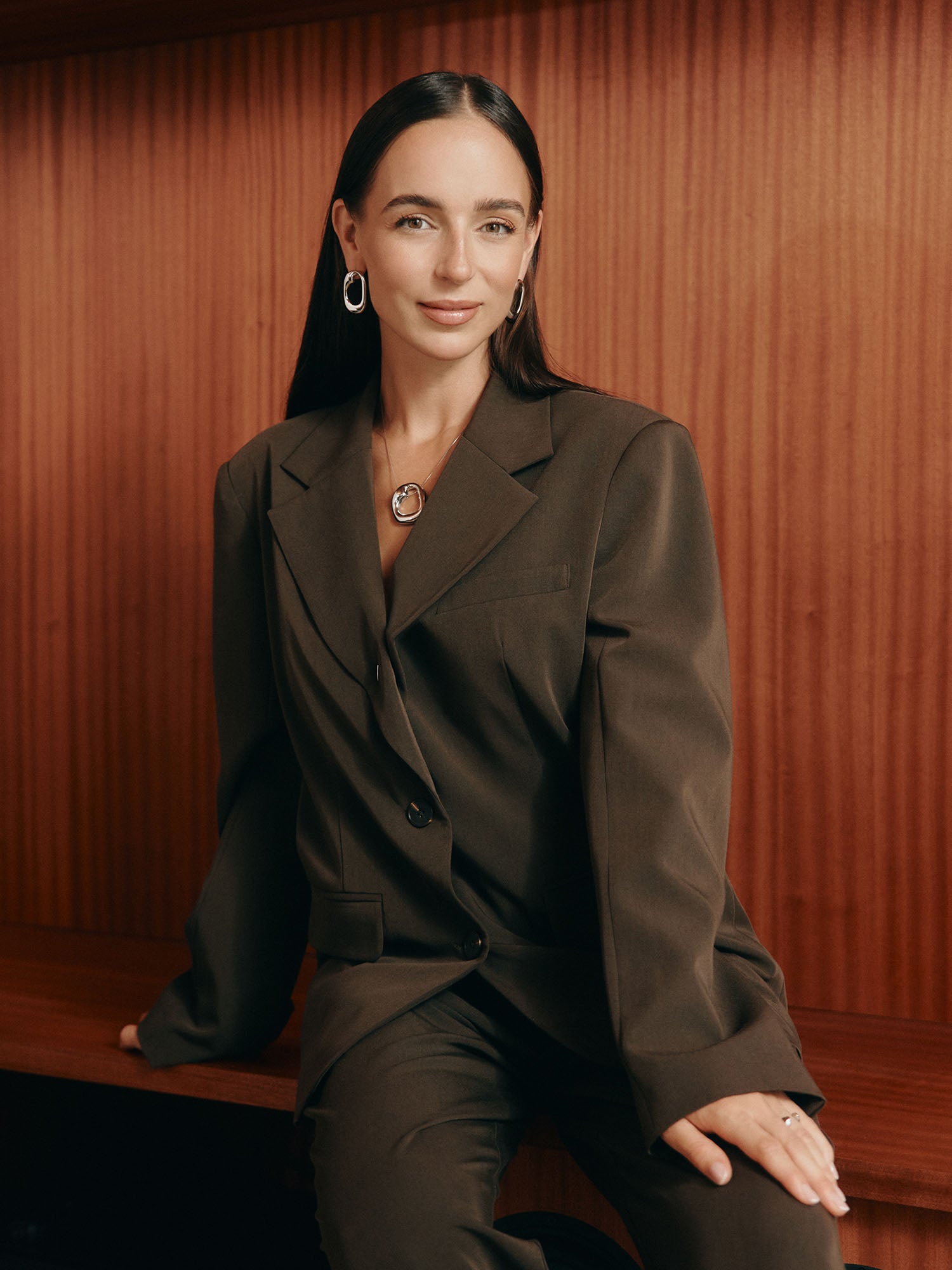 Eine Frau mit langen dunklen Haaren, in einem braunen Anzug und Silberschmuck, sitzt auf einer Holzbank vor Holzpaneelen und lächelt sanft - ihr Stil ist so raffiniert wie die LeGer x Purelei Athina Pants von LeGer by Lena Gercke.