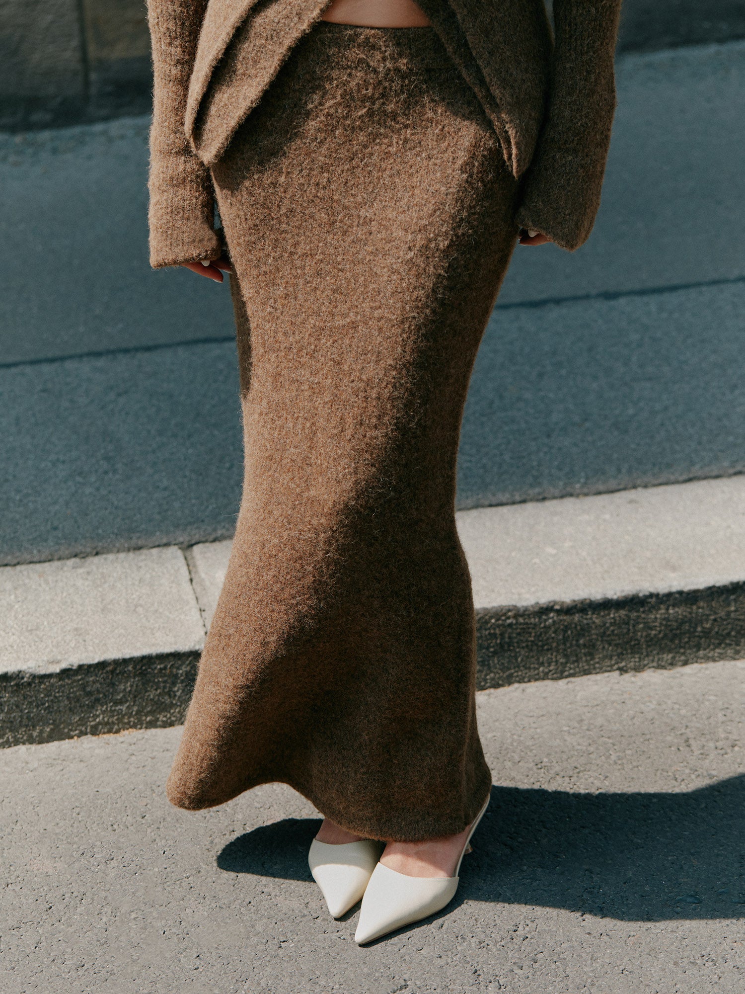 Eine Person trägt den Frederike Rock von LeGer by Lena Gercke mit einem passenden braunen Strickoberteil und weißen spitzen Absätzen, während sie mit den Händen an der Seite auf einem Gehweg steht. Das Foto ist oberhalb der Schultern abgeschnitten.