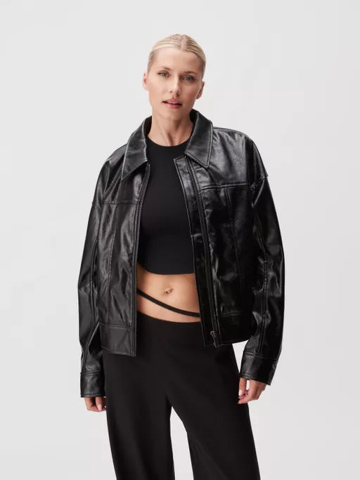Eine Person, die eine schwarze Kunstlederjacke über einem schwarzen bauchfreien Oberteil und einer schwarzen Hose trägt, steht vor einem schlichten weißen Hintergrund. Ihre Haare sind zurückgekämmt und sie hat einen neutralen Gesichtsausdruck.