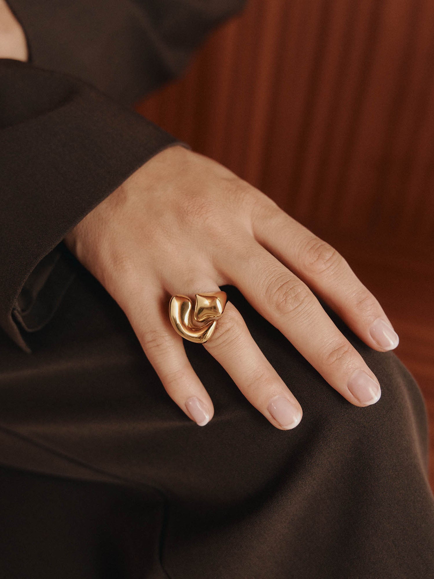 Eine Hand mit gepflegten Nägeln ruht auf dunkelbraunem Stoff und trägt den LeGer x Purelei - Fluid Forms Ring von LeGer by Lena Gercke - einen kühnen, skulpturalen Ring am Mittelfinger.