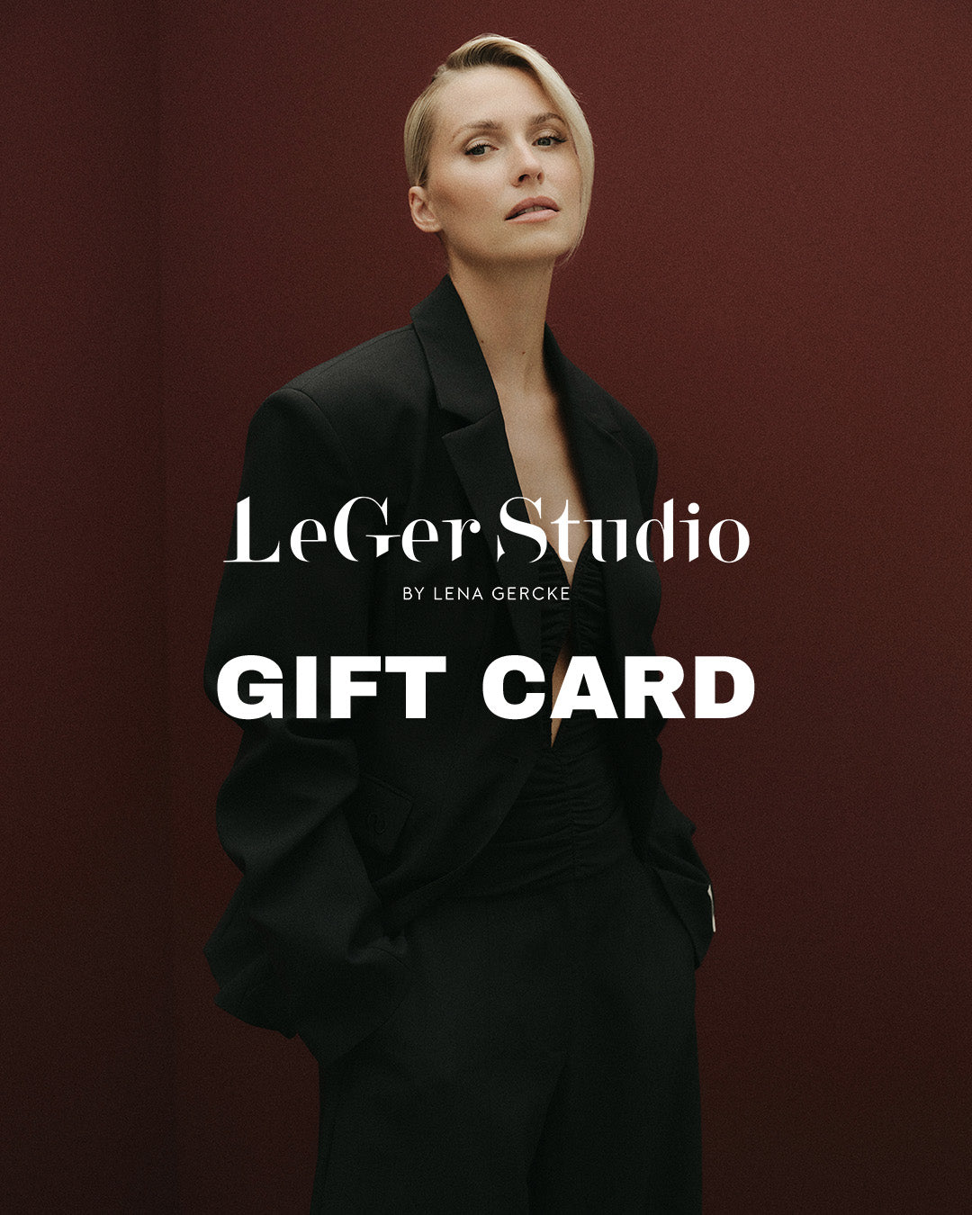 Eine Frau in einem schwarzen Blazer steht vor einem dunkelroten Hintergrund einer LeGer by Lena Gercke-Geschenkkarte, auf dem die Schriftzüge „LeGer Studio BY LENA GERCKE“ und „GIFT CARD“ zu sehen sind.