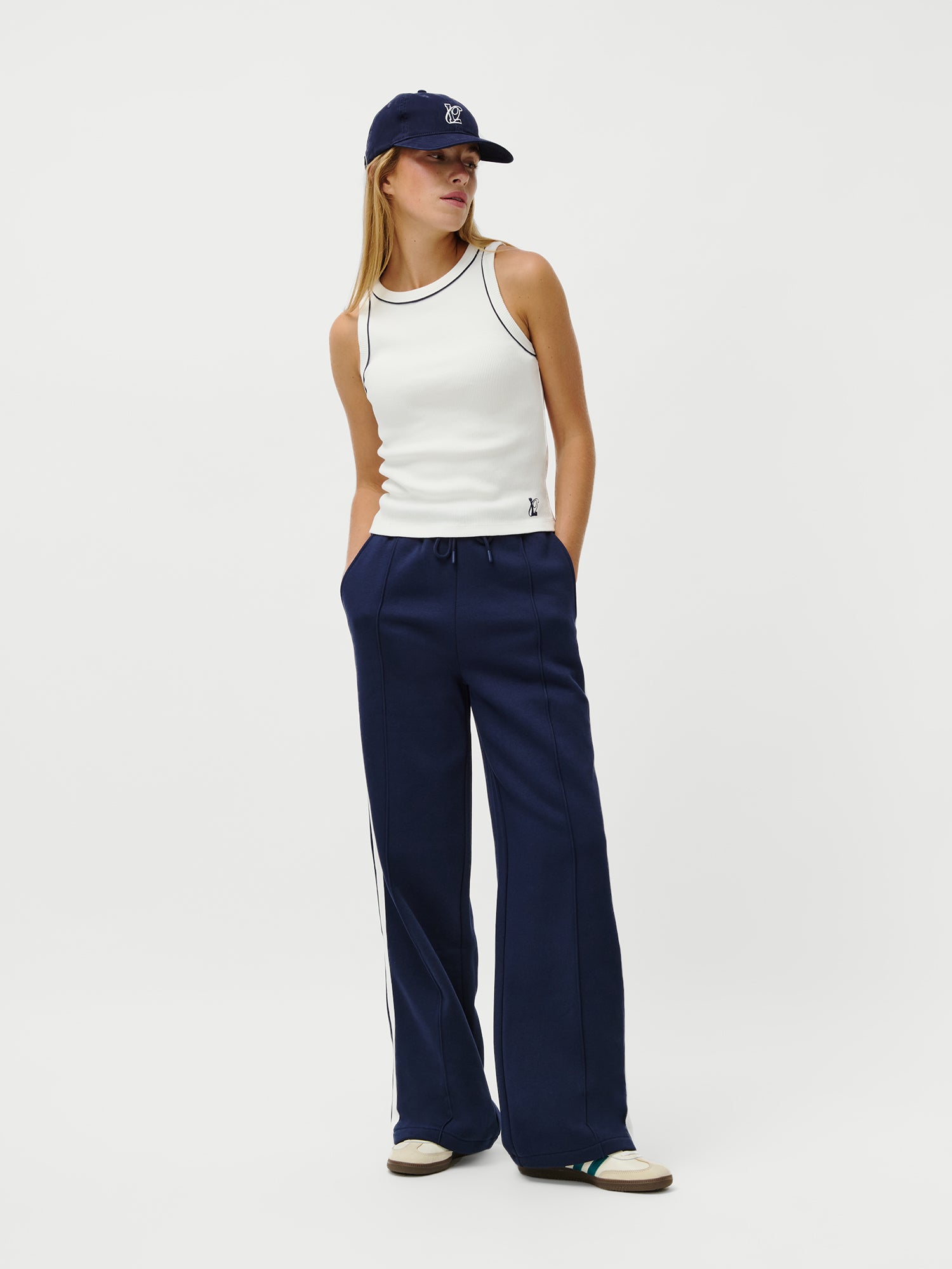 Eine Frau steht vor einem weißen Hintergrund und trägt das schmal geschnittene LeGer x Oh April - Dira Top von LeGer by Lena Gercke, kombiniert mit einer navyfarbenen Kappe, einer navyfarbenen Hose mit weitem Bein und weißem Seitenstreifen und beigen Sneakers. Ihre Hände stecken in den Taschen.