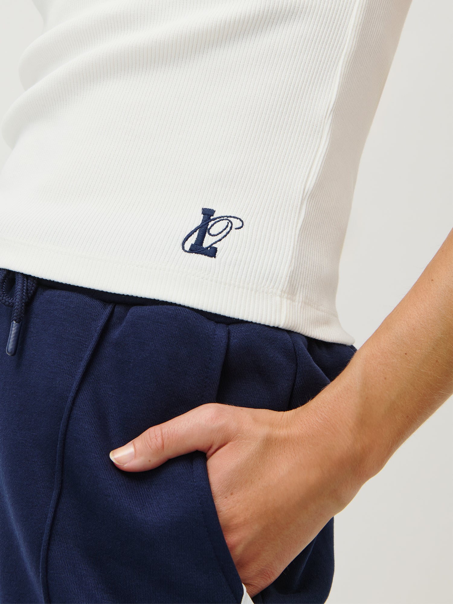 Eine Person trägt das LeGer x Oh April - Dira Top von LeGer by Lena Gercke, ein weißes, ärmelloses Slim Fit Rib-Tanktop mit kleinem "L" am Saum und kombiniert es mit navyblauen Sweatpants vor schlichtem Hintergrund.
