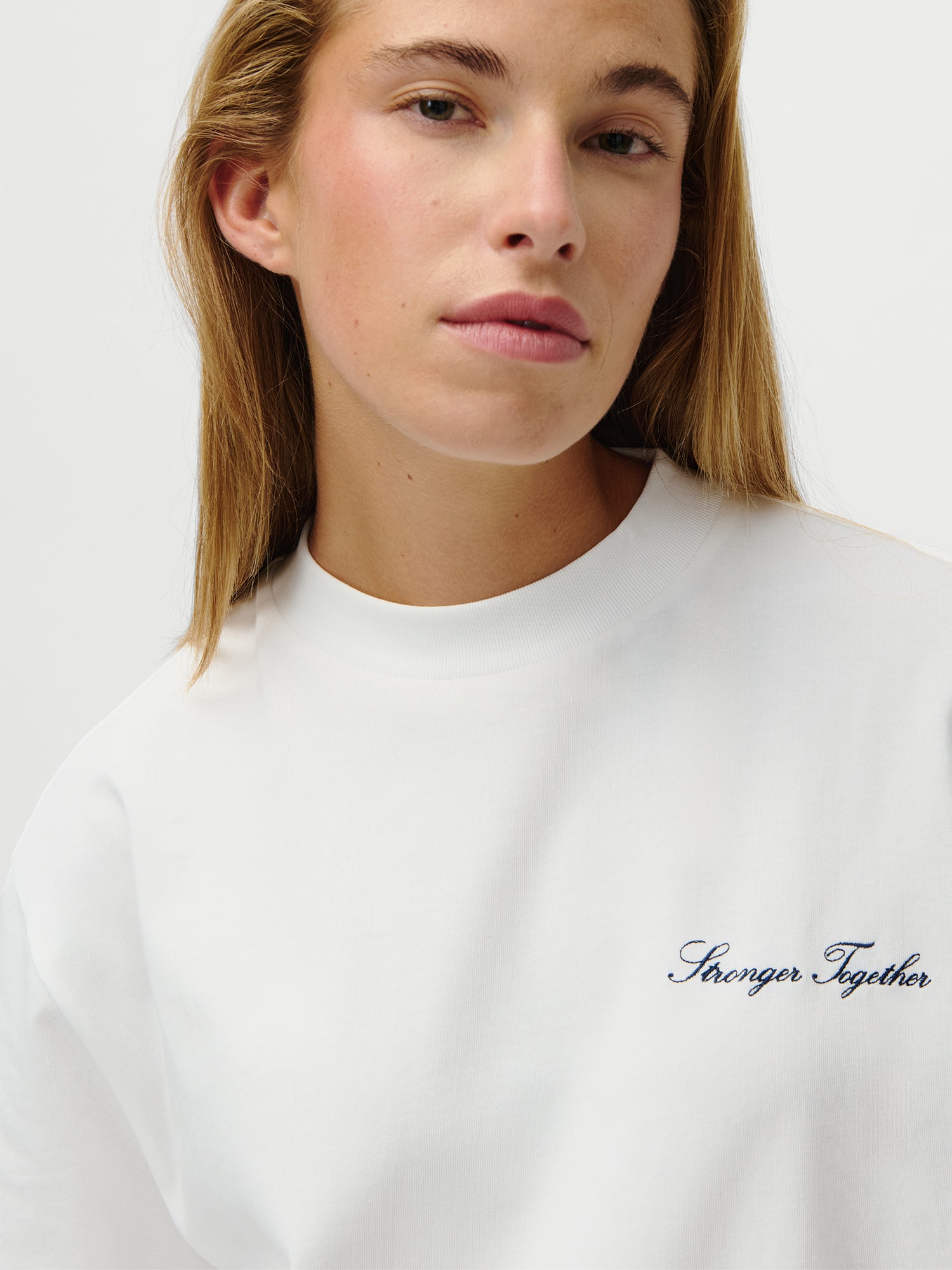 Eine Person mit langen blonden Haaren trägt das LeGer x Oh April - Amara T-Shirt Stronger by LeGer by Lena Gercke-weiße, schmal geschnittene Viertelarm Shirt in normaler Länge mit "Stronger Together" Stickerei auf der Brust und schlichtem, hellem Hintergrund.
