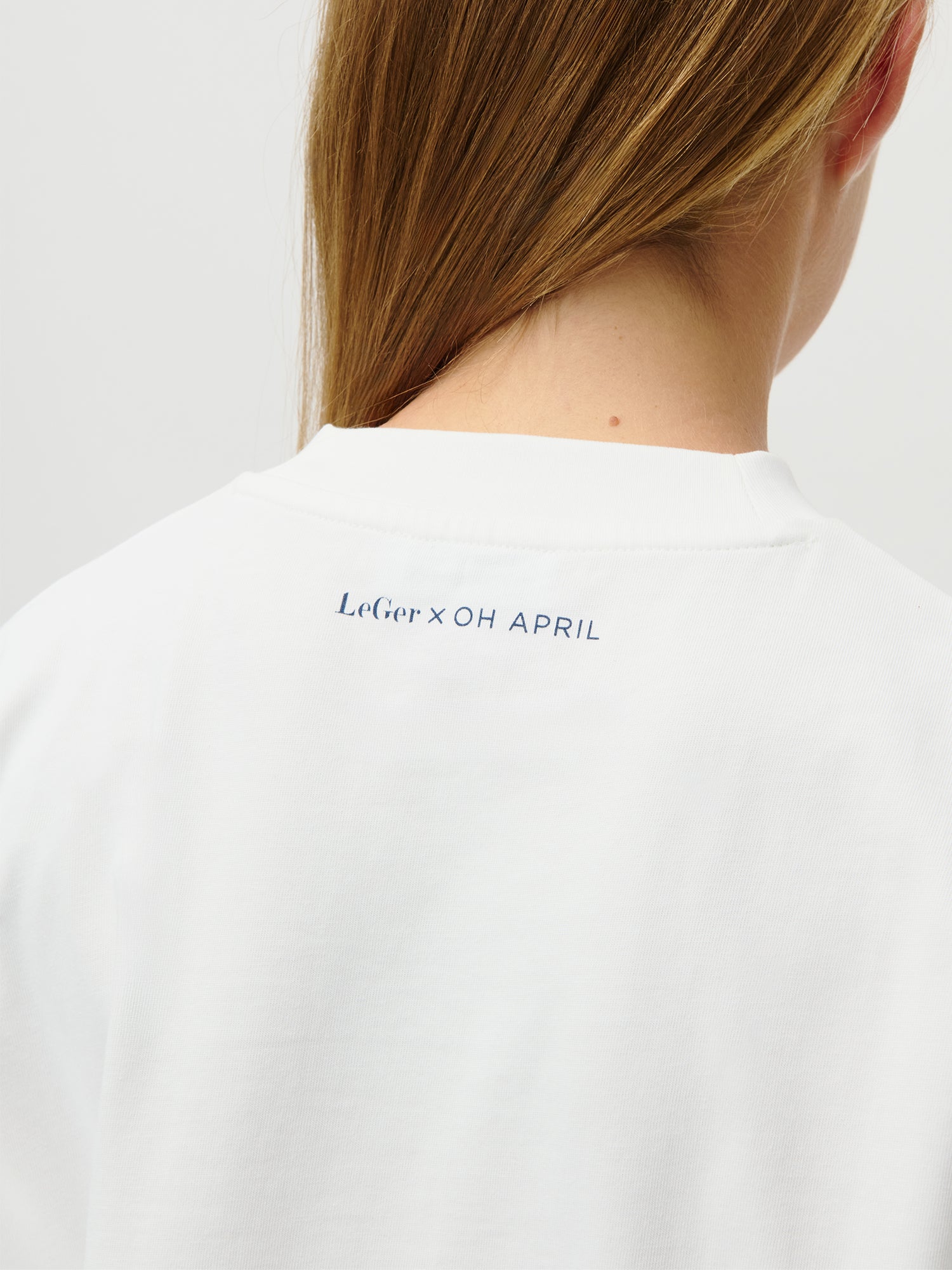 Eine Nahaufnahme des oberen Rückens einer Person im LeGer by Lena Gercke "LeGer x Oh April - Amara T-Shirt Stronger": weiß, schmale Passform, normale Länge, mit kleinem blauen "LeGer x OH APRIL"-Schriftzug am Kragen. Blonde Haare fallen locker über eine Schulter.