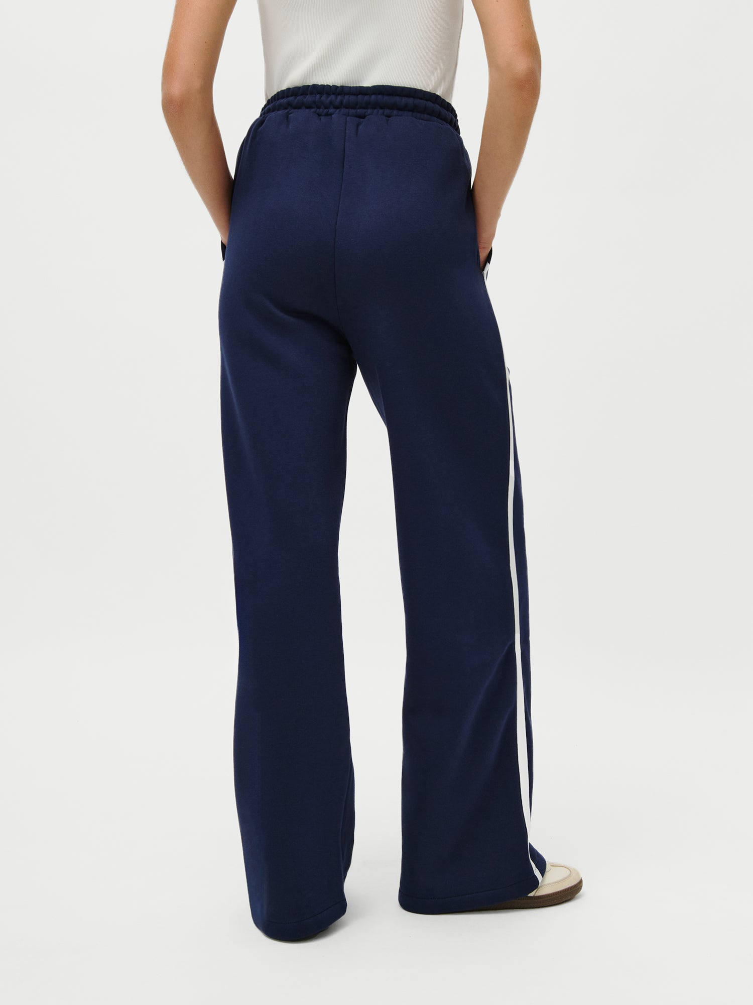 Eine Person (1,73m) trägt den LeGer x Oh April - Joan Jogger von LeGer by Lena Gercke in navy mit weißem Seitenstreifen und weißen Schuhen, steht mit den Händen in den Taschen von der Kamera abgewandt vor einem schlichten Hintergrund.