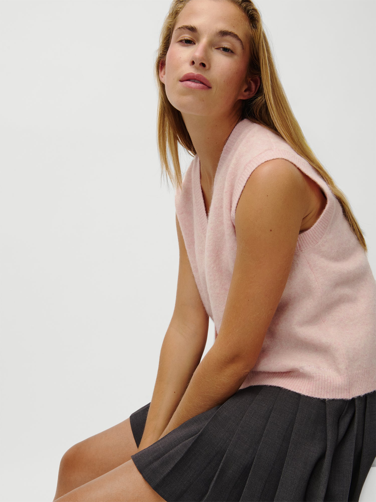 Eine Frau mit langen blonden Haaren trägt die LeGer x Oh April - Amira Knit Vest von LeGer by Lena Gercke, einen hellrosa ärmellosen Pullover in Größe 36 mit Passform normal und einen dunkelgrauen Faltenrock. Sie sitzt und schaut mit neutralem Blick nach oben. Größe des Modells: 1,73 m.