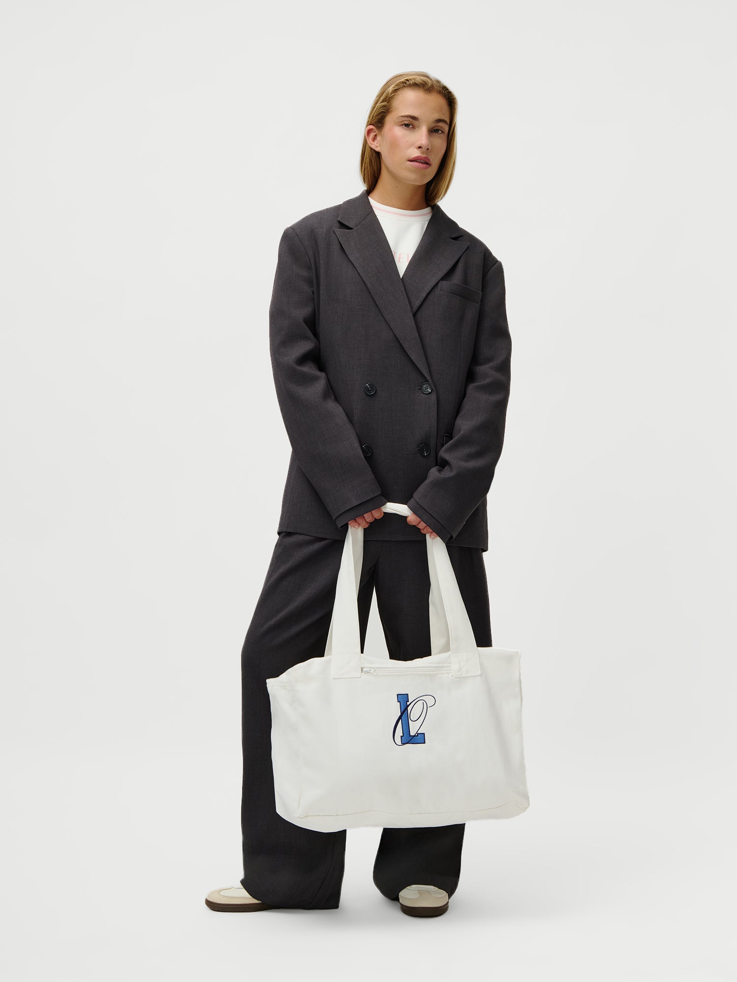 Eine Person mit langen Haaren trägt einen lockeren dunklen Anzug, ein weißes Hemd und graue Schuhe (Größe 36) und steht vor einem weißen Hintergrund. Das Model (1,73m) trägt den LeGer x Oh April - Anni Shopper von LeGer by Lena Gercke, eine große weiße Tasche mit blau-schwarzem Monogramm-Design.