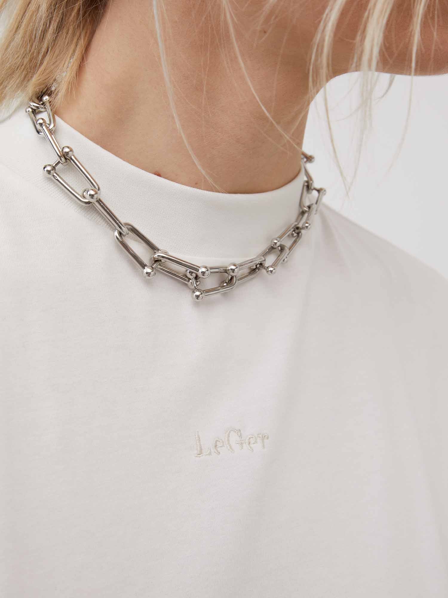 Nahaufnahme einer Person, die ein weißes Hemd „Irina“ von LeGer von Lena Gercke trägt, das mit „Le Ger“ bestickt ist. Sie trägt eine klobige silberne Halskette und ihr Haar fällt teilweise über ihre Schulter. Beachten Sie das Viertelarm-Design des Hemdes, das zu seinem einzigartigen Stil beiträgt.