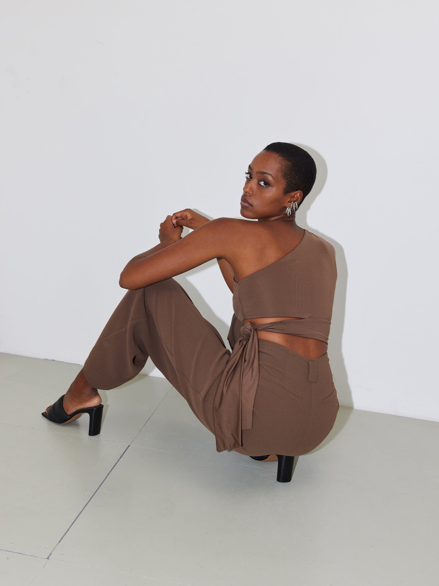 Eine Person mit Kurzschnitt-Frisur trägt ein stylisches Top „Leia“ von LeGer by Lena Gercke mit One-Shoulder-Design in Braun, dazu passende schmale braune Hosen. Sie hockt mit einem angehobenen Knie und blickt über die Schulter in die Kamera. Sie trägt schwarze Stöckelschuhe und der Hintergrund ist schlicht weiß.