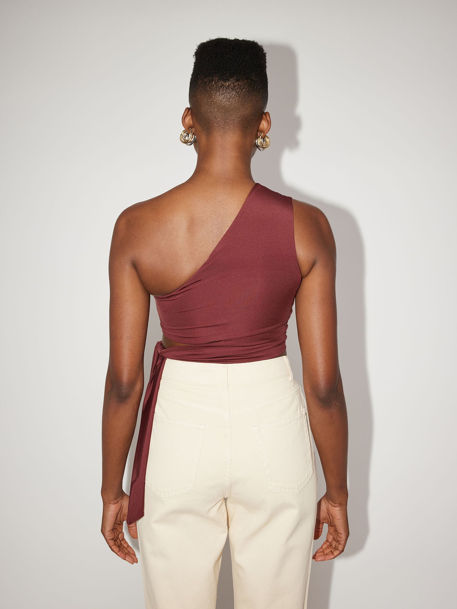 Rückansicht einer Person mit kurzen Haaren, die das schmale, burgunderfarbene Leia Top von LeGer by Lena Gercke und weiße Hosen trägt. Das ärmellose Top wird auf der rechten Seite elegant gebunden, vor einem schlichten hellgrauen Hintergrund.