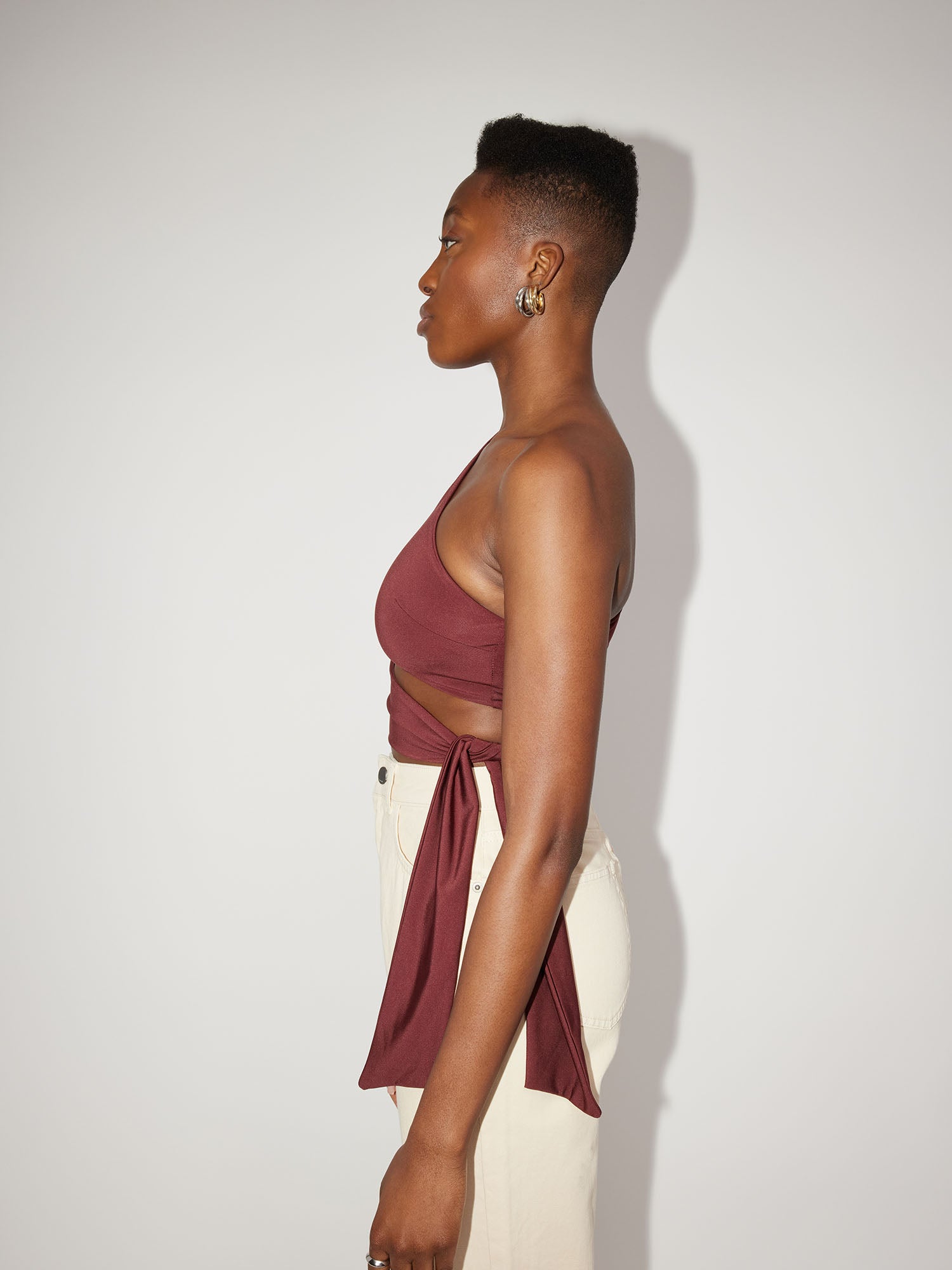 Eine Person steht im Profil vor einem schlichten Hintergrund und präsentiert das Leia Top von LeGer by Lena Gercke in Kombination mit hellen Hosen. Mit kurzen Haaren im kurzen Schnitt und Creolen als Accessoire ist ihr Look sowohl elegant als auch modern.