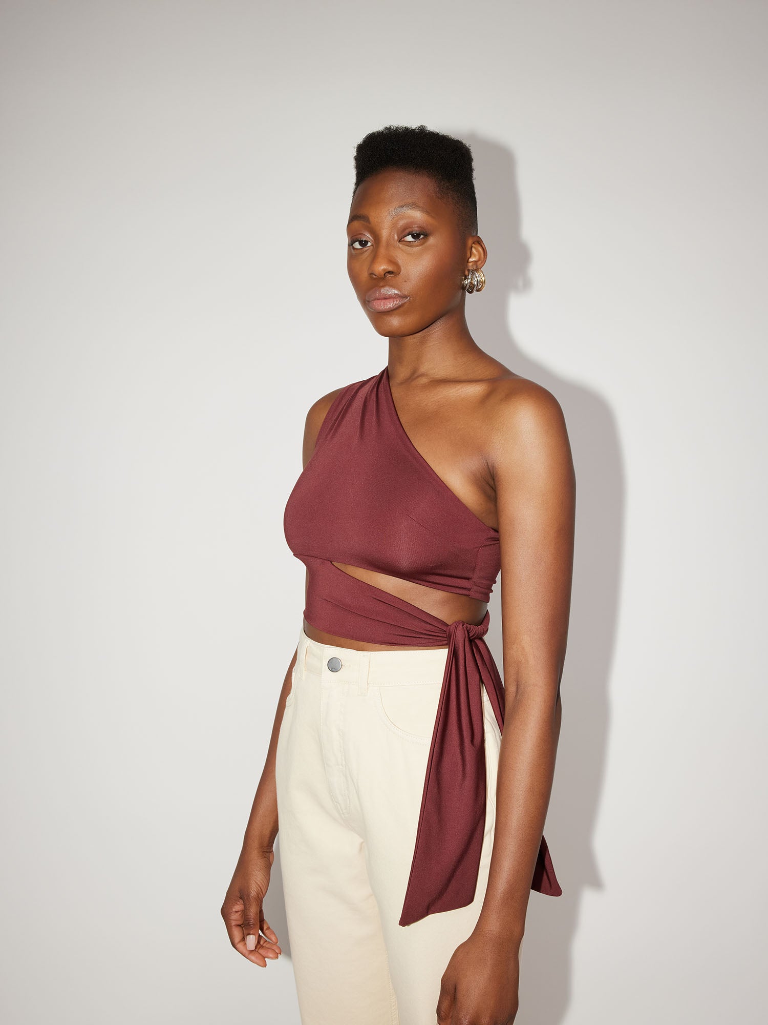 Eine Person steht selbstbewusst vor einem schlichten Hintergrund und präsentiert das burgunderfarbene Leia Top von LeGer by Lena Gercke – ein ärmelloses One-Shoulder-Kleid mit asymmetrischem Design – kombiniert mit beigen Hosen. Der neutrale Hintergrund und die sanfte Beleuchtung betonen die schlanke Silhouette des Tops.