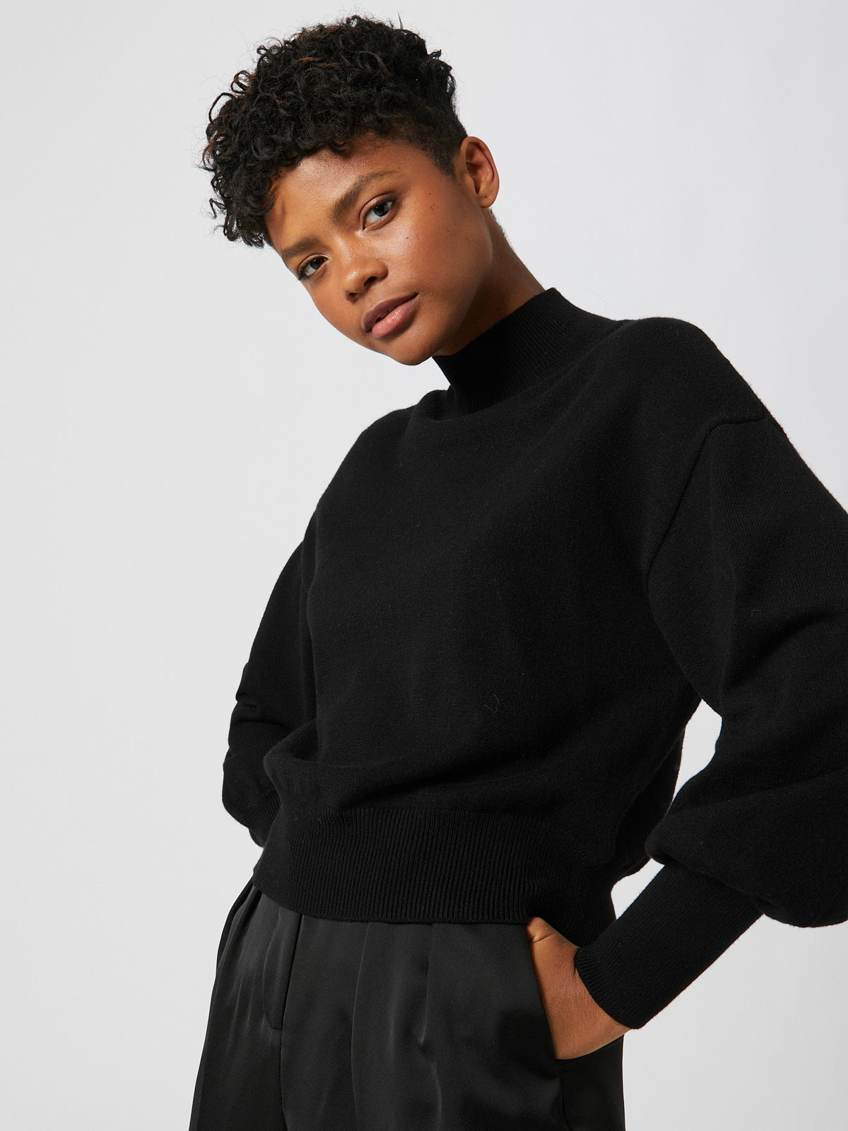 Eine Person mit kurzen Locken trägt einen schwarzen „Penelope“-Pullover von LeGer by Lena Gercke in normaler Länge, dazu eine schwarze Hose, und steht in einer entspannten und selbstbewussten Pose vor einem schlichten, hellen Hintergrund.