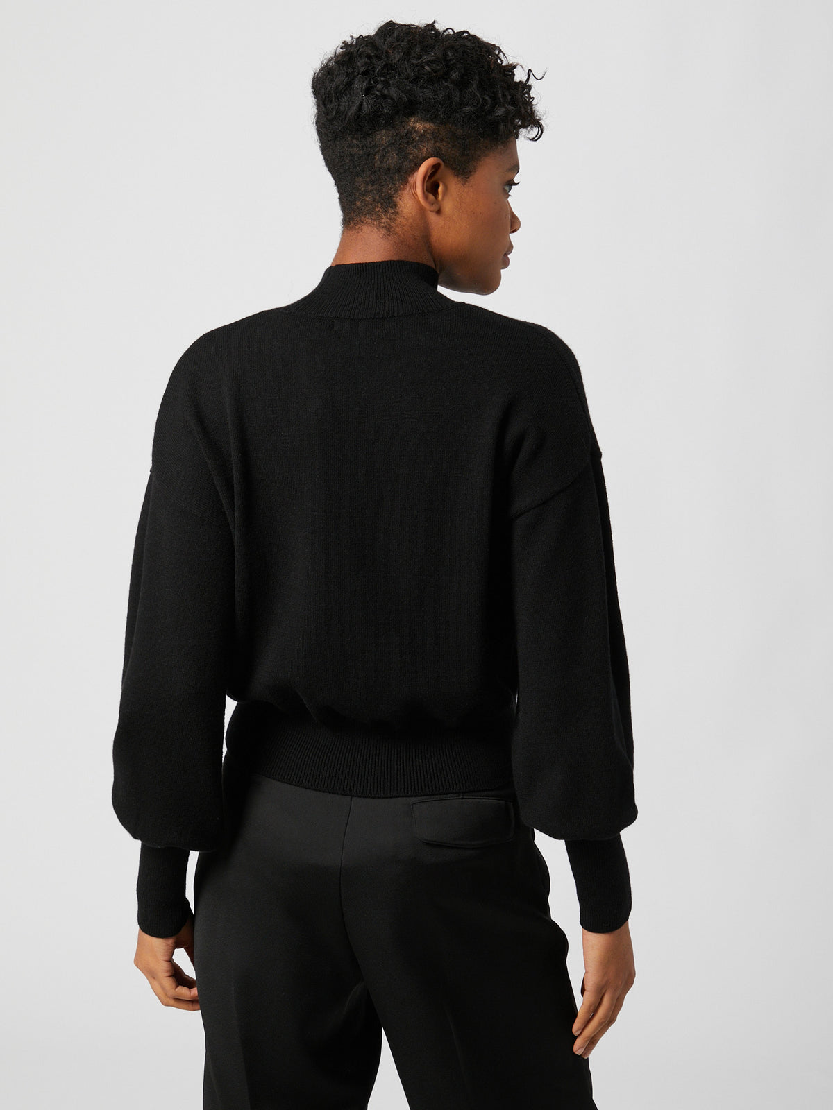 Eine Person mit kurzen Locken, die den schwarzen Langarm-Pullover „Penelope“ von LeGer by Lena Gercke und eine schwarze Hose in normaler Länge trägt, steht mit dem Rücken zur Kamera vor einem schlichten, hellen Hintergrund und präsentiert dabei eine lässige, moderne Passform.