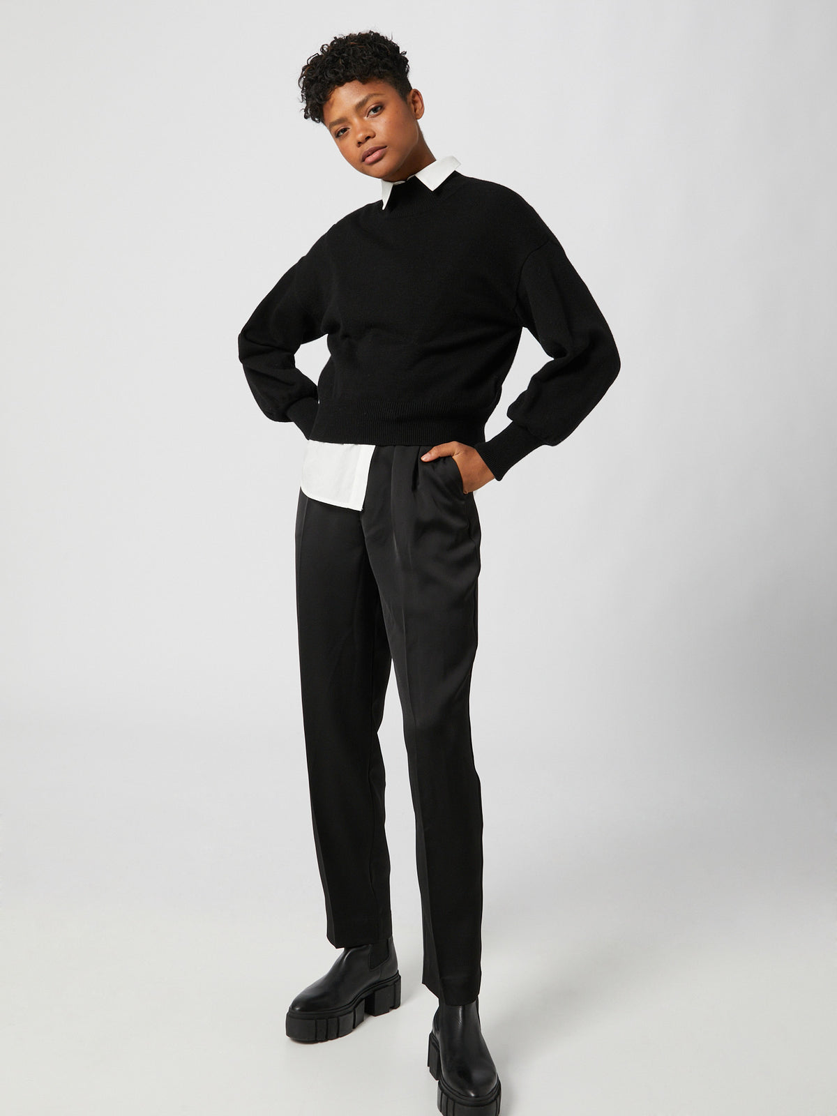 Eine Person steht vor einem schlichten Hintergrund und trägt den schwarzen Pullover „Penelope“ von LeGer by Lena Gercke über einem weißen Hemd mit Kragen, dazu eine schwarze Hose in normaler Länge und klobige schwarze Stiefel. Mit den Händen in den Taschen und einem neutralen Gesichtsausdruck wird die lockere Passform des Outfits besonders hervorgehoben.