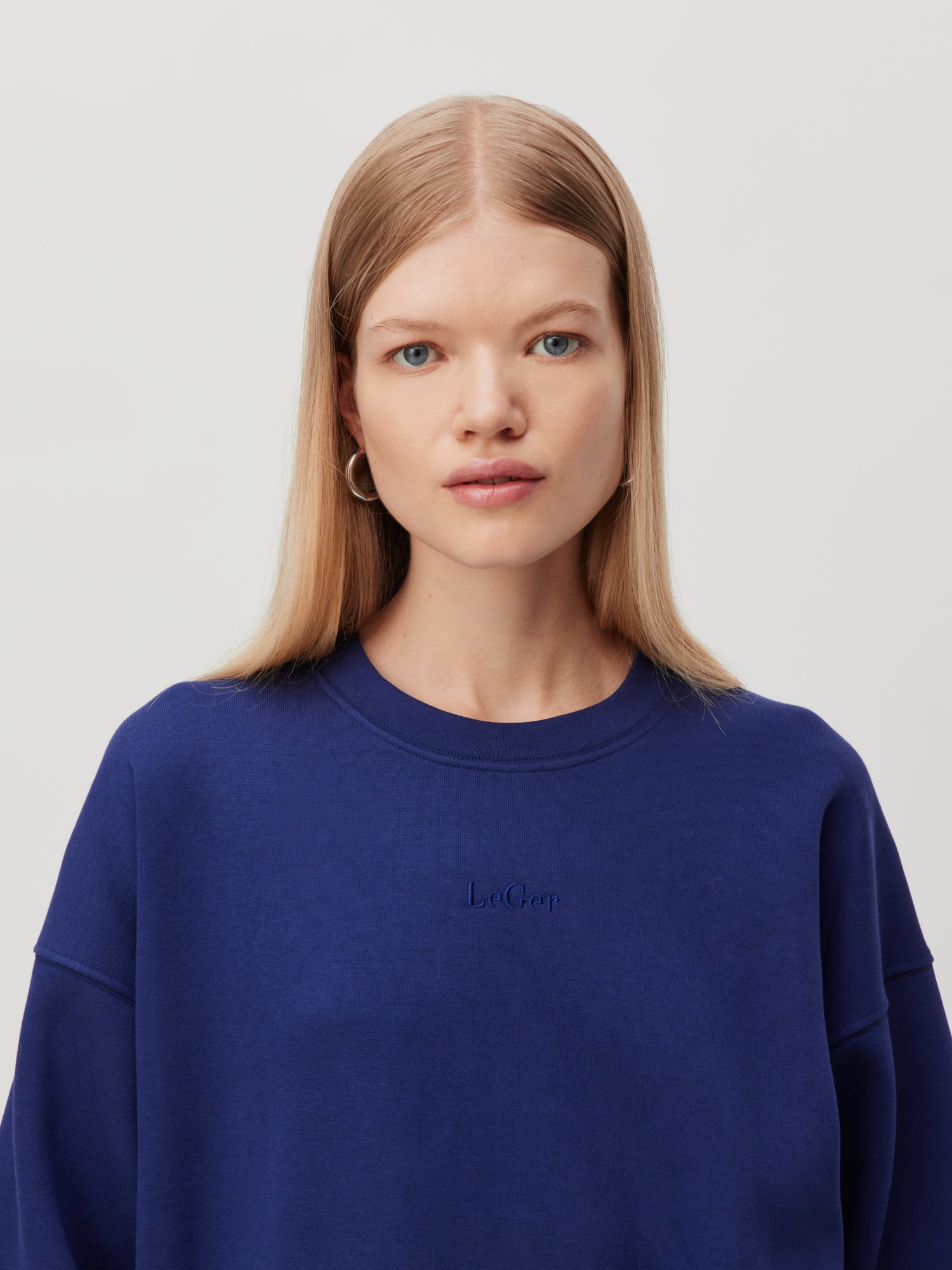 Eine Person mit langen glatten blonden Haaren ist von den Schultern aufwärts zu sehen, nach vorne gerichtet. Sie trägt ein schlichtes blaues LeGer by Lena Gercke Sweatshirt 'Indra' in normaler Länge mit der Aufschrift "Le Gap" auf der Brust. Der Hintergrund ist schlicht und hell.
