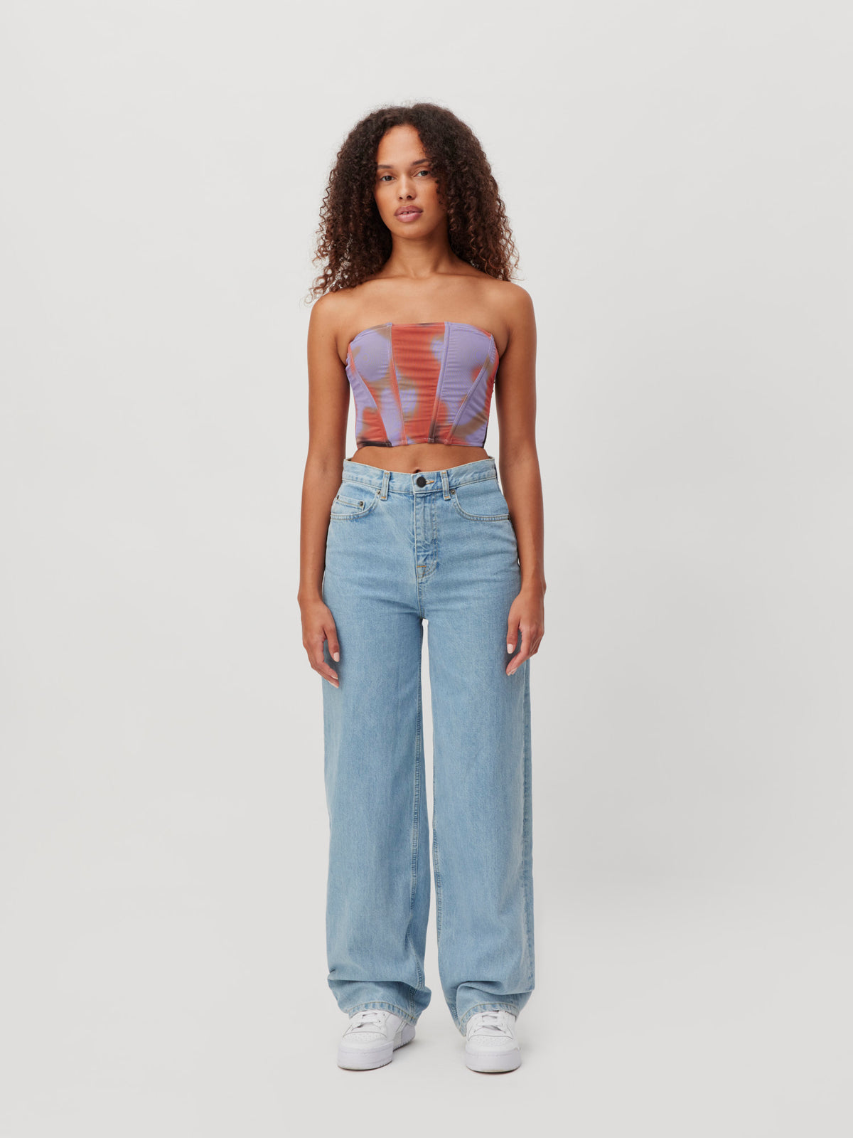 Eine Frau, die ein trägerloses Korsett-Top mit Tie-Dye-Muster, eine hellblaue, weit geschnittene Jeanshose „Carla“ von LeGer by Lena Gercke sowie weiße Sneaker trägt.