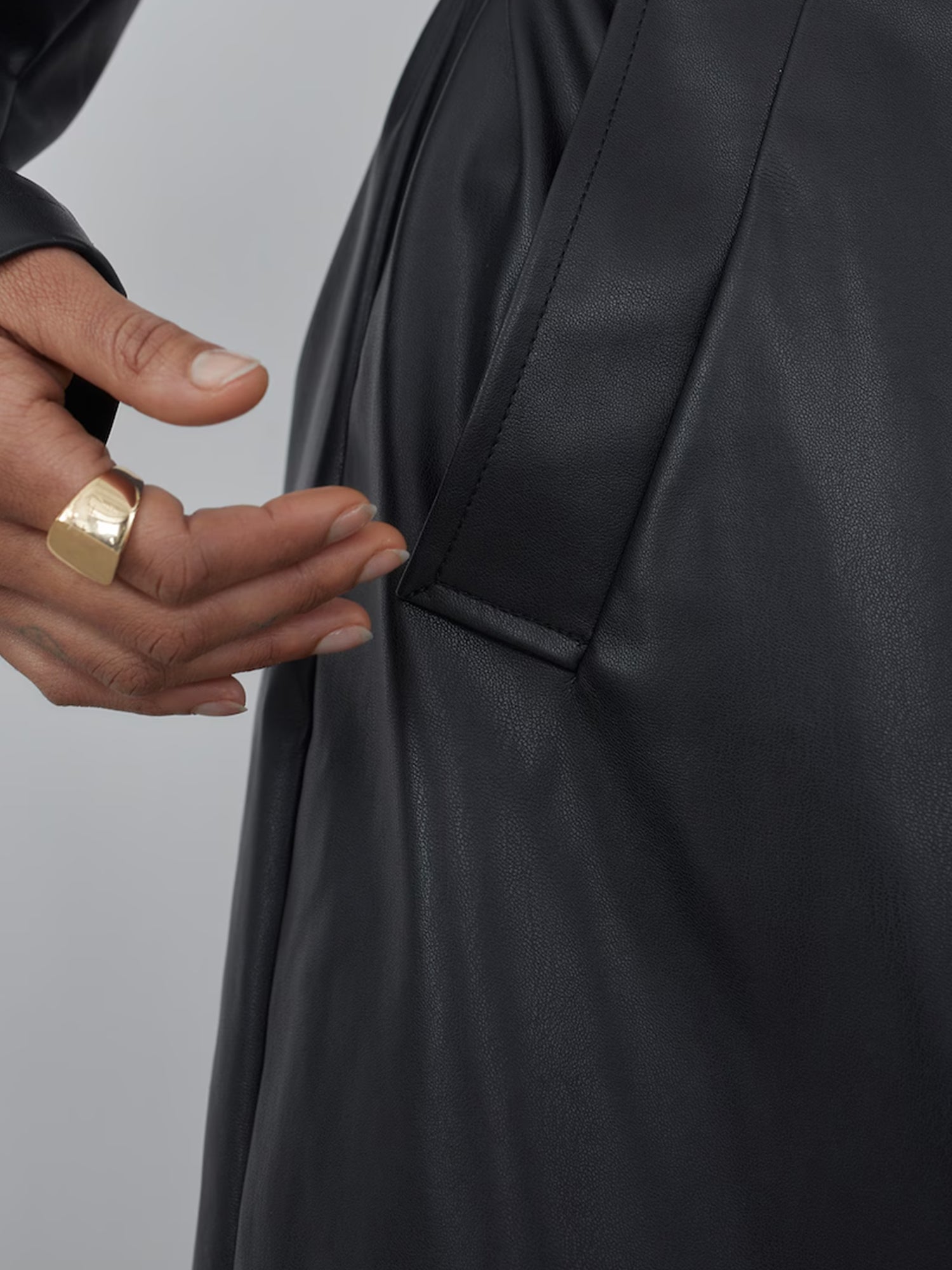 Eine Nahaufnahme einer menschlichen Hand, die mit einem goldenen Ring geschmückt ist und nach der Seitentasche einer Marla-Jacke von LeGer by Lena Gercke greift. Die Textur des schwarzen Leders und die Nähte um die Tasche herum sind deutlich zu erkennen und heben die tadellose Passform hervor.