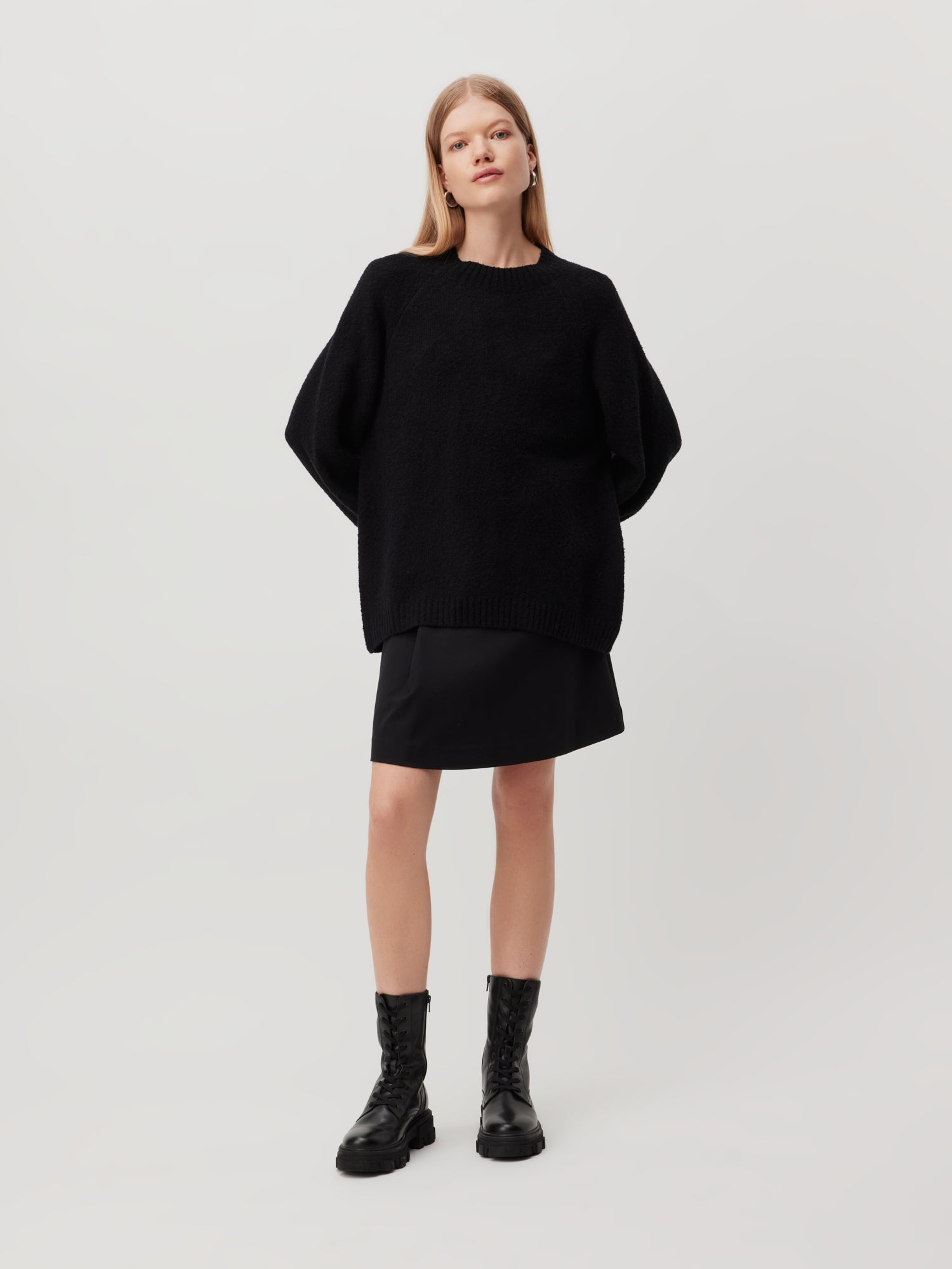 Vor einem schlichten, hellen Hintergrund steht eine Person, die einen schwarzen, übergroßen Pullover „Elmira“ von LeGer by Lena Gercke trägt, dazu einen schwarzen Rock und schwarze Schnürstiefel. Sie hat lange, glatte Haare und blickt direkt in die Kamera.