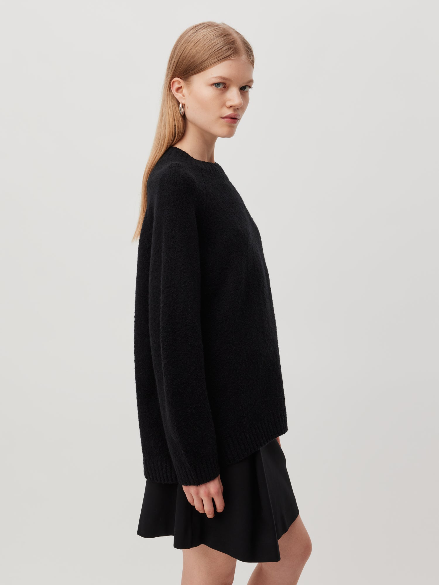 Eine Person mit langen, glatten Haaren steht seitlich und trägt einen locker sitzenden schwarzen Pullover namens Pullover ‚Elmira‘ von LeGer by Lena Gercke und einen schwarzen Rock von Normale Länge. Der schlichte, helle Hintergrund unterstreicht das lässige, minimalistische Outfit. Der Ausdruck der Person ist neutral.